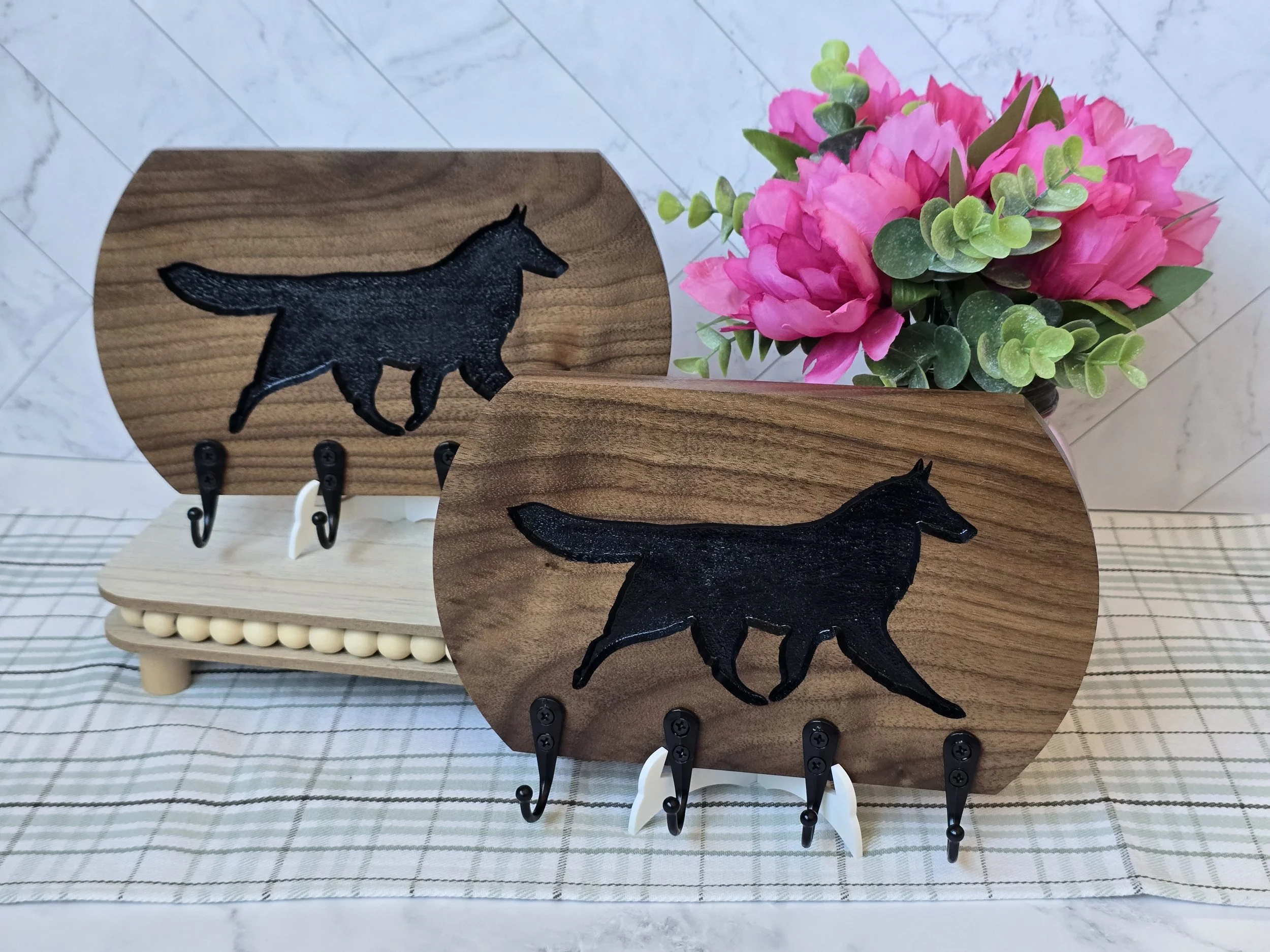 Decorative leash holder featuring a black silhouette of a Belgian Tervuren, displayed on a wooden board, with pink flowers and green foliage arranged behind it.