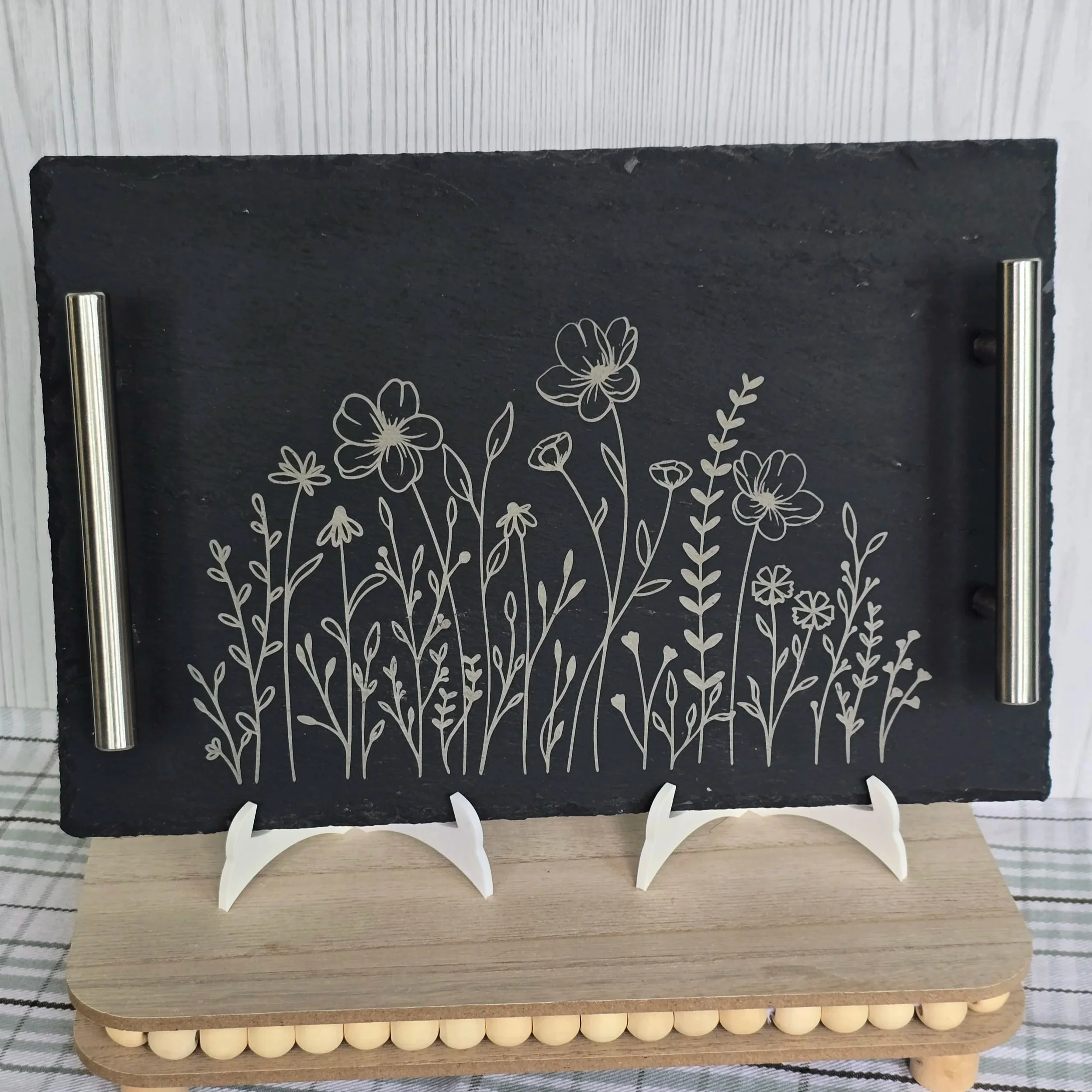 A decorative blackboard with white floral drawings, mounted on a wooden stand with white legs, placed on a light-colored surface.