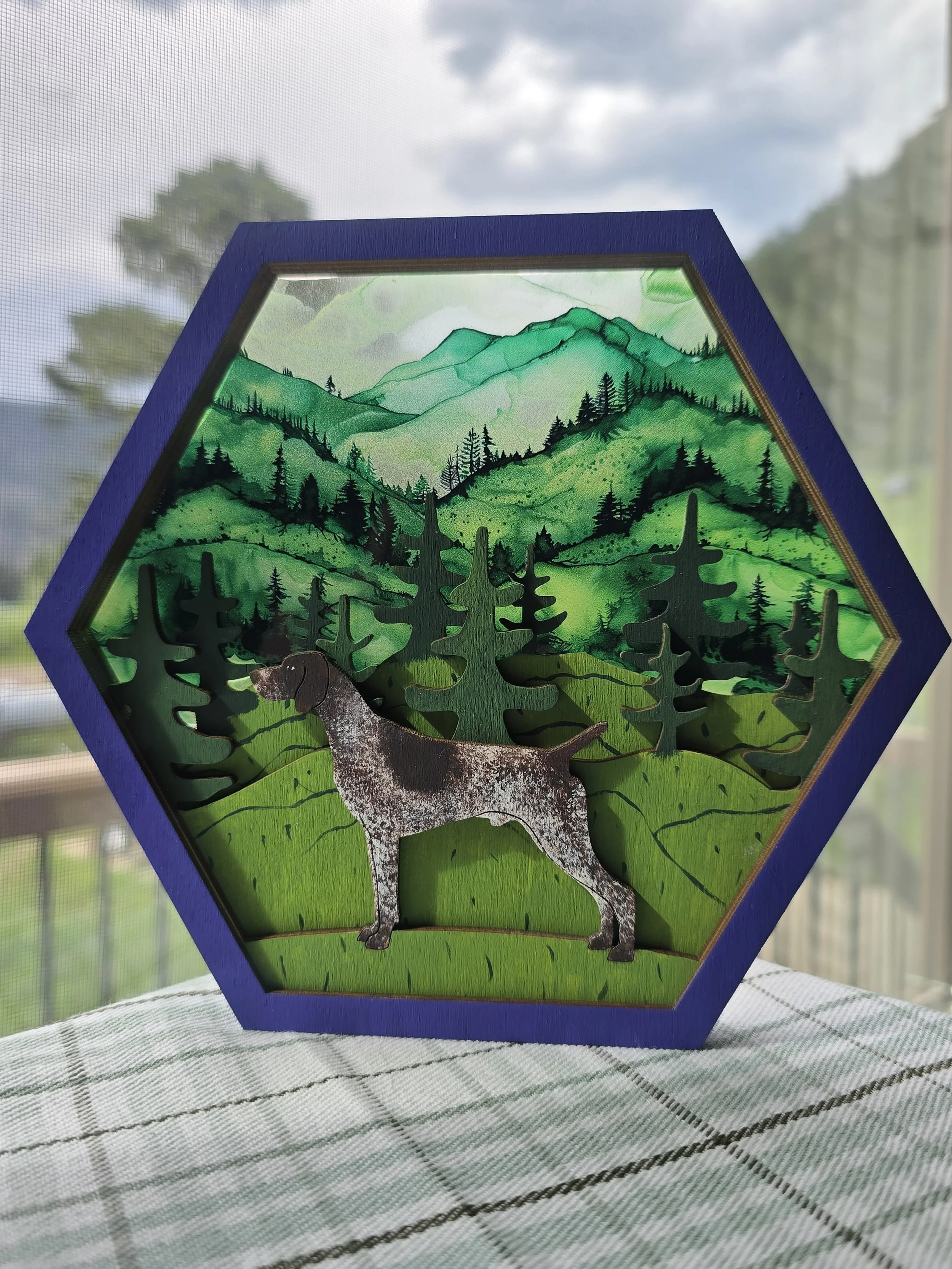A hand painted artwork of a German Shorthaired Pointer with background of mountainous landscape and pine trees, with a patterened acrylic background, framed with a purple border, placed on a white and green checkered cloth.