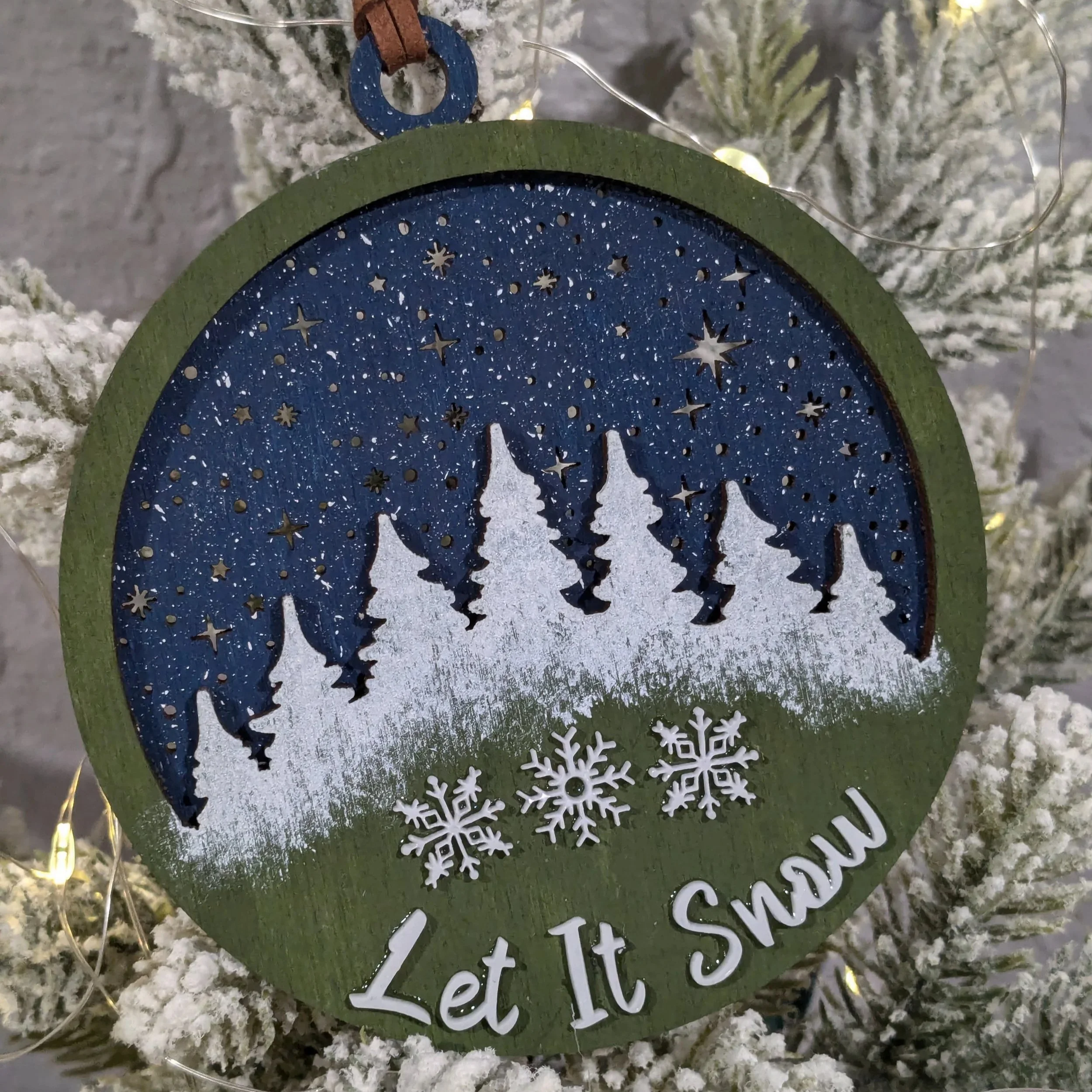 Close-up of a Christmas ornament hanging on a snow-covered Christmas tree. The ornament is circular with a green border and a blue interior featuring small star and snowflake cutouts, revealing a white silhouette of a Christmas tree. The background shows white artificial snow and some fuzzy white branches.