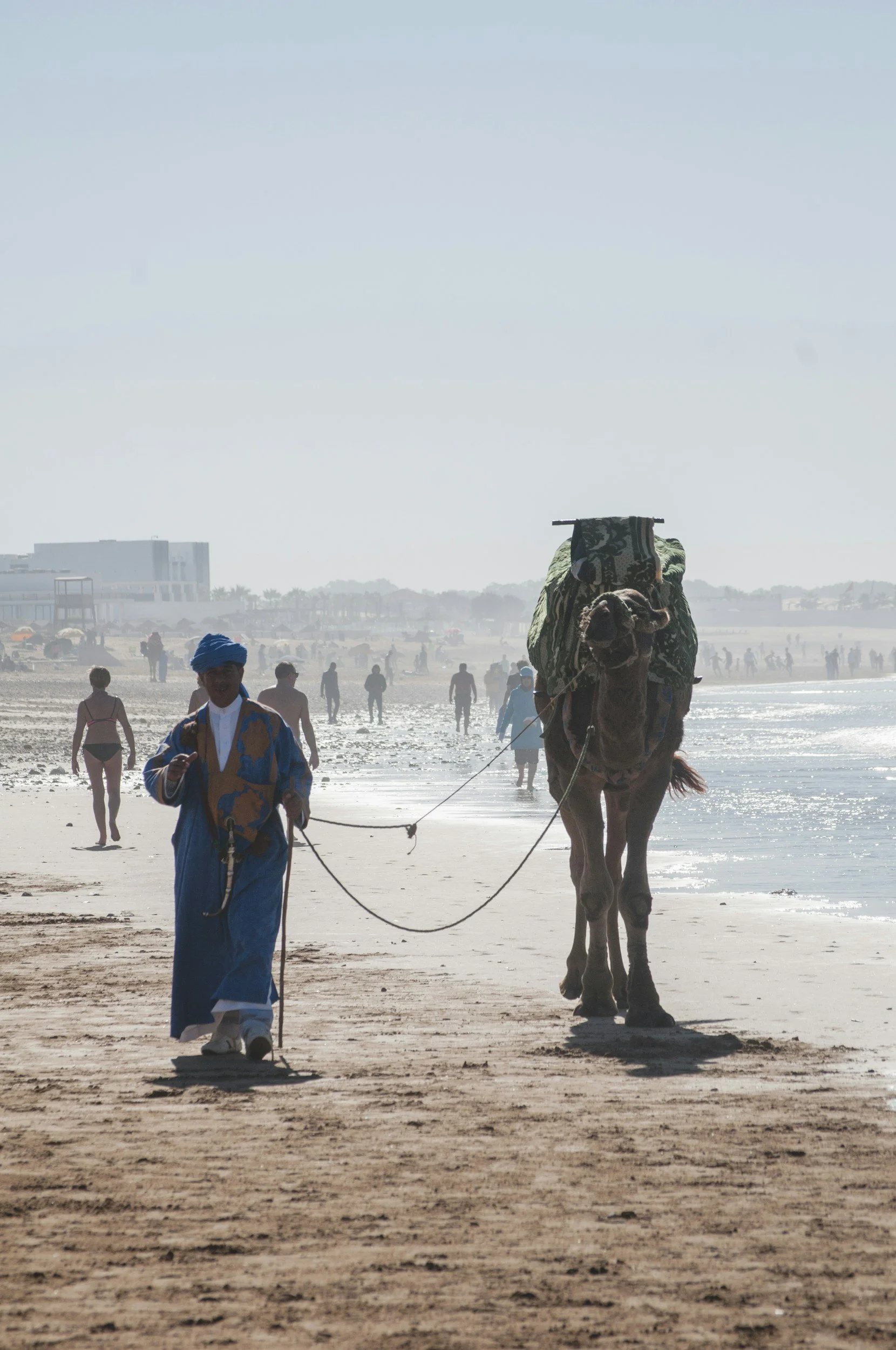 Une personne en vêtement traditionnel marche sur la plage à Agadir avec un chameau chargé, en arrière-plan d'autres personnes se promènent sur la plage et dans l'eau.