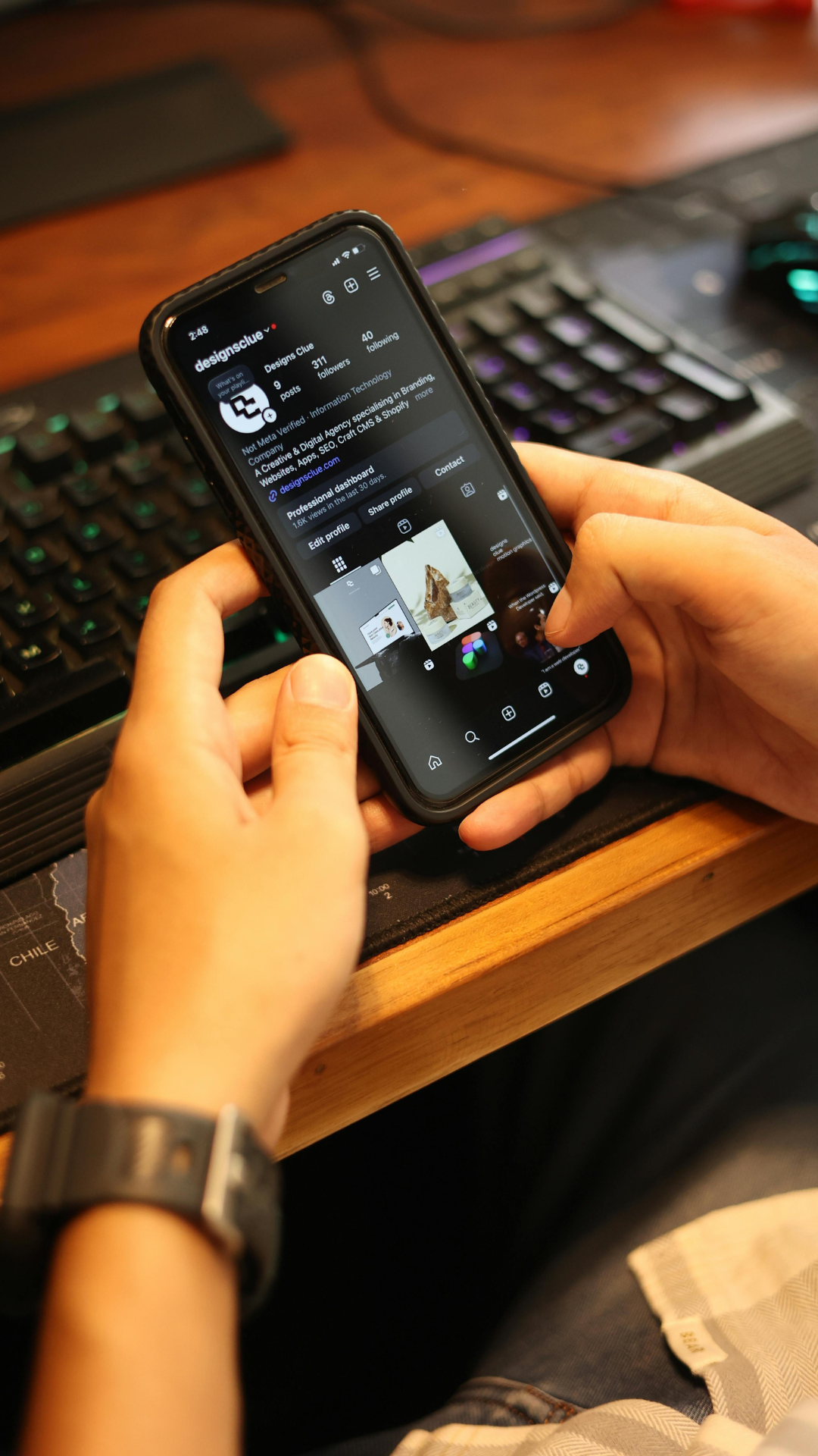 Close-up of a person holding a smartphone displaying an Instagram profile, with a keyboard partially visible on a wooden desk in the background.