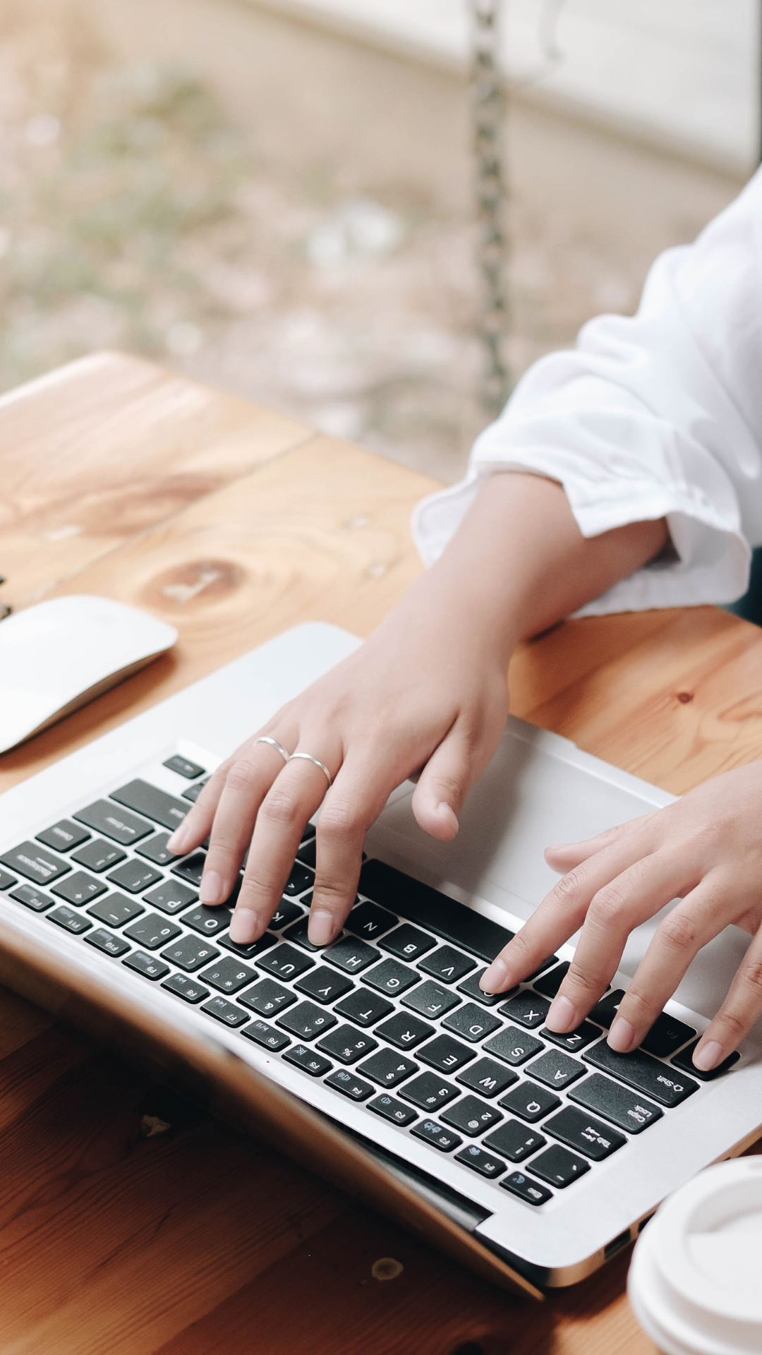 Person typing on a silver laptop with black keys, sitting at a wooden table, near a white computer mouse and a to-go coffee cup.