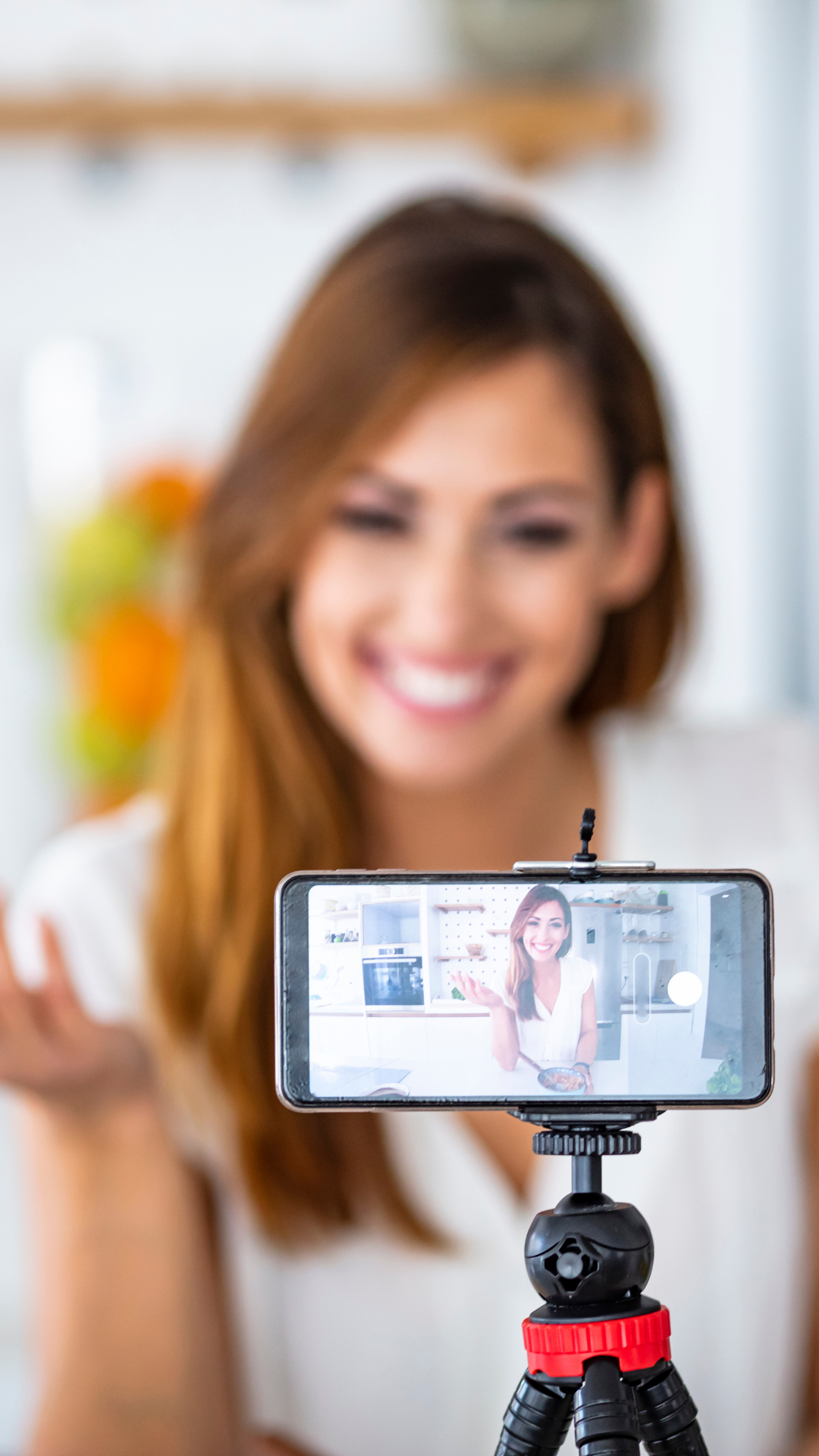 A woman with brown hair smiling at a camera while taking a selfie with her smartphone on a tripod. The phone screen shows her smiling face and the background of a modern kitchen.