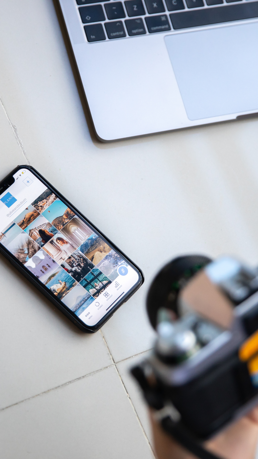 Close-up of a smartphone on a white surface, displaying a grid of travel photos, with a laptop keyboard partly visible in the background and blurred objects in the foreground.