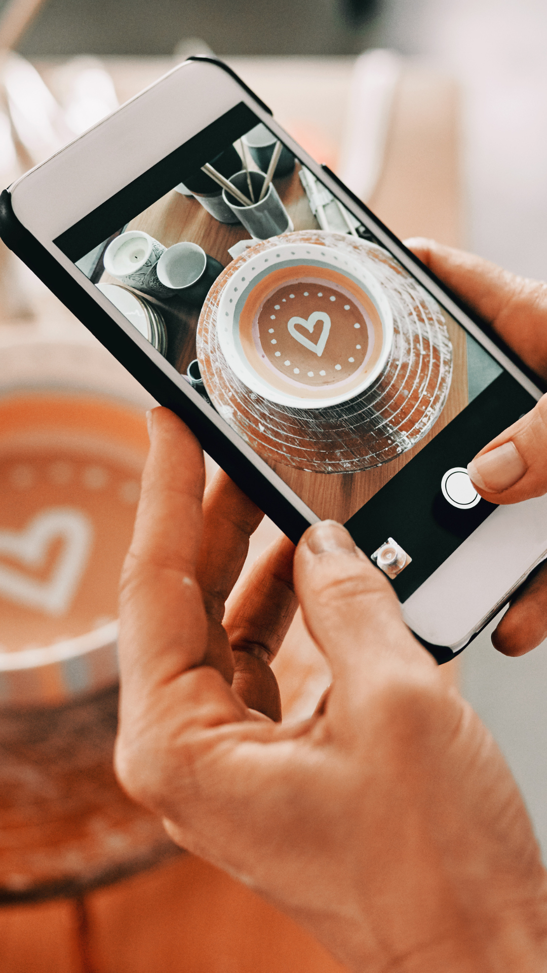 Person taking a photo of a latte with a heart design on top using a smartphone at a cafe.