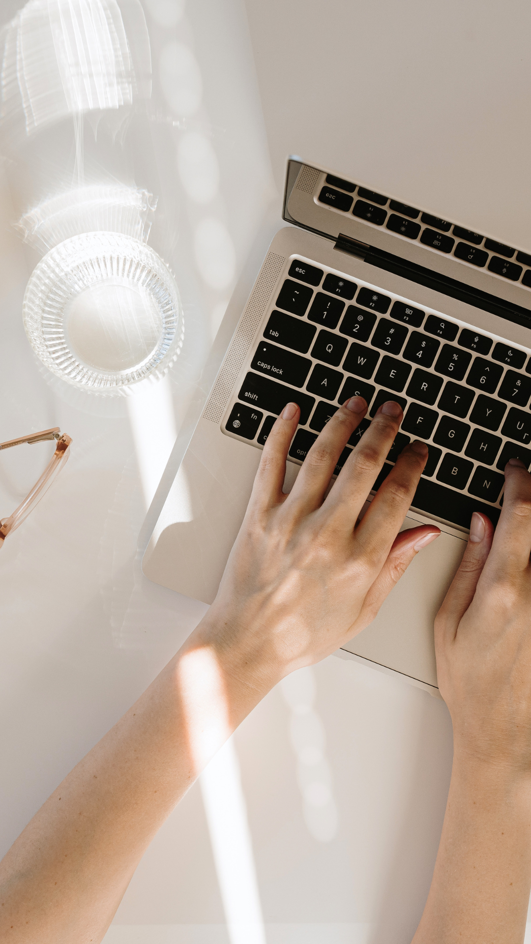 Person typing on a silver laptop keyboard with sunlight creating patterns on a white surface, alongside glasses and a glass dish.
