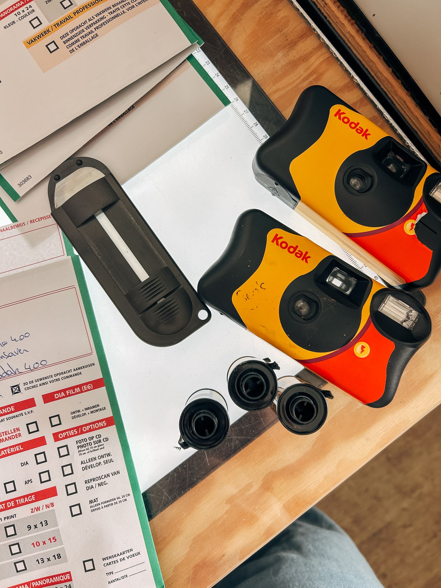Flat lay of two Kodak disposable cameras, three film canisters, a film cutter, and photo processing forms on a table with a ruler edge.