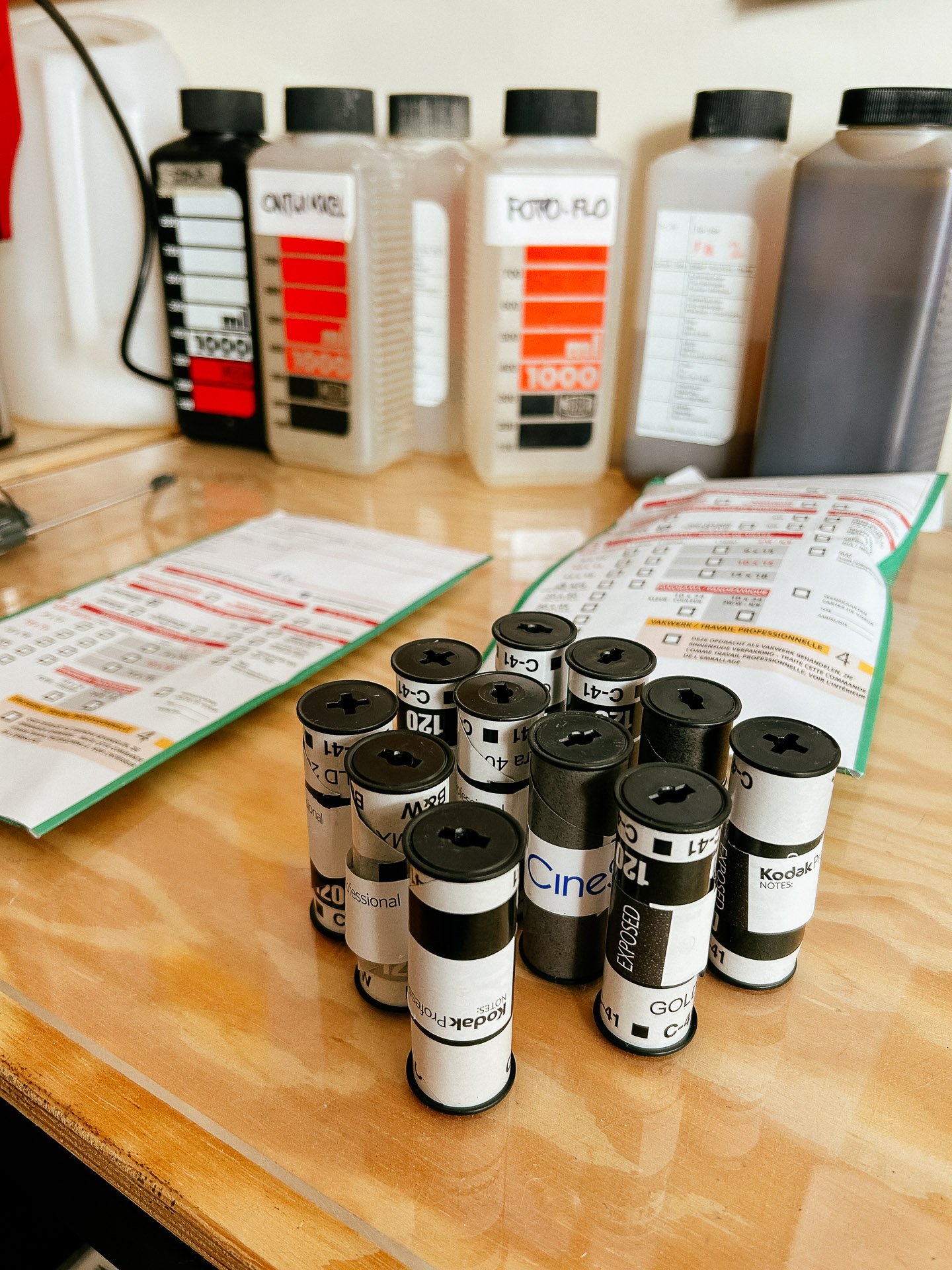 A collection of photographic film canisters on a wooden table, with chemical bottles labeled "C41," "FOTO-FLO," and others in the background. Instruction sheets are also visible.