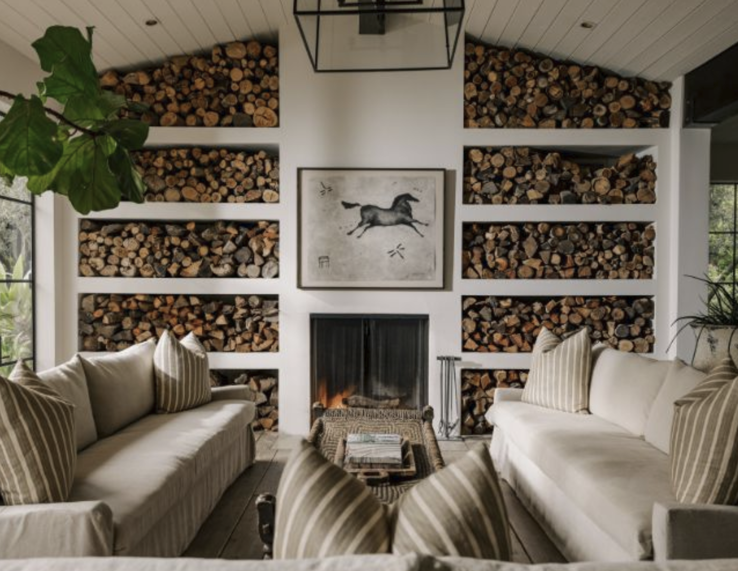 Living room with white walls and a white wood-paneled ceiling. Two white sofas with striped pillows face each other around a wooden coffee table. Behind the sofas, a white wall features built-in shelves filled with neatly stacked firewood, a framed horse artwork above a fireplace, and a large green plant on the left. Large windows on both sides let in natural light.