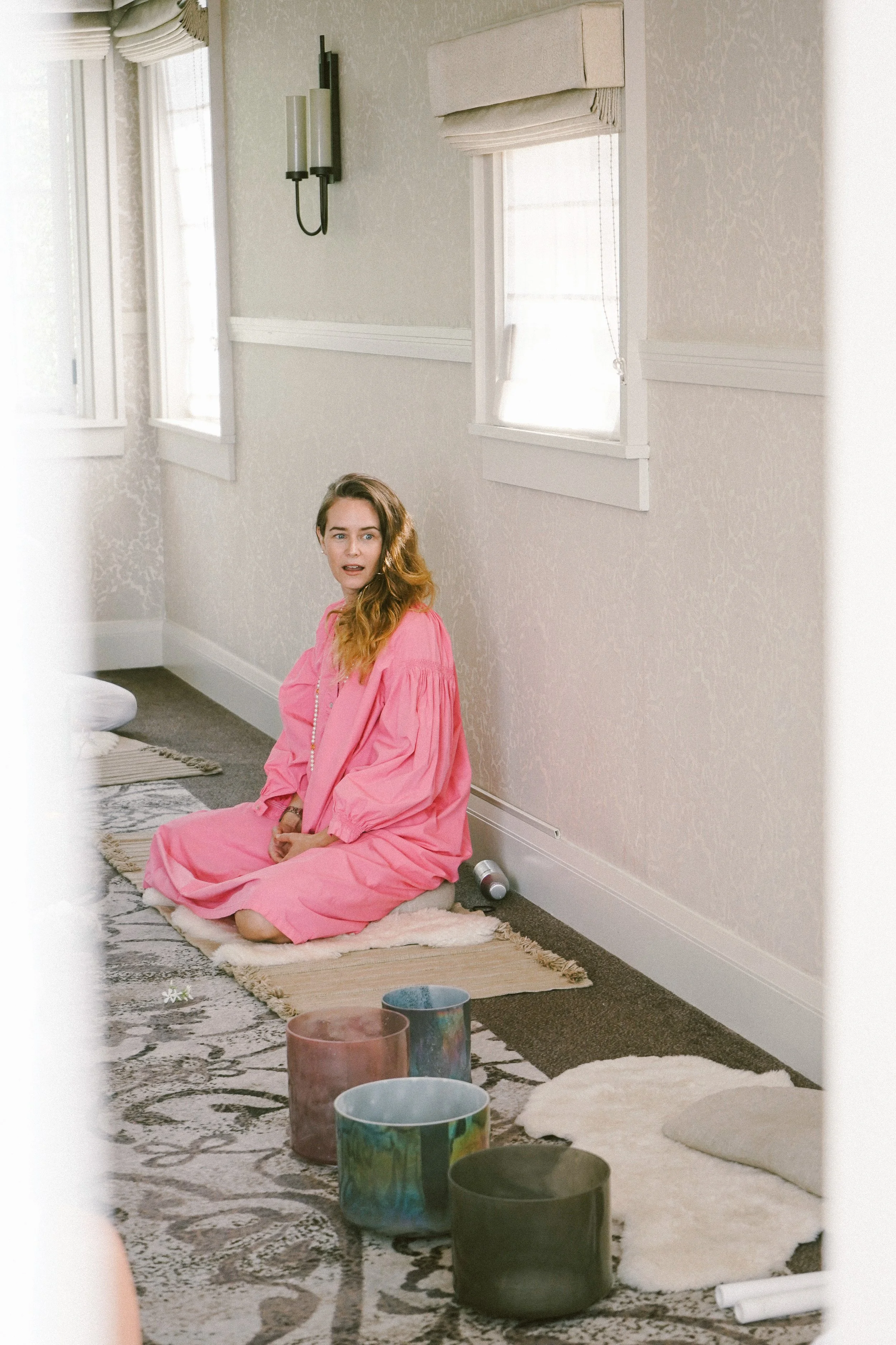 Person in a pink dress sitting on a rug in a peaceful room with light-colored walls, near a window. Several colorful crystal singing bowls are placed on the floor next to them.