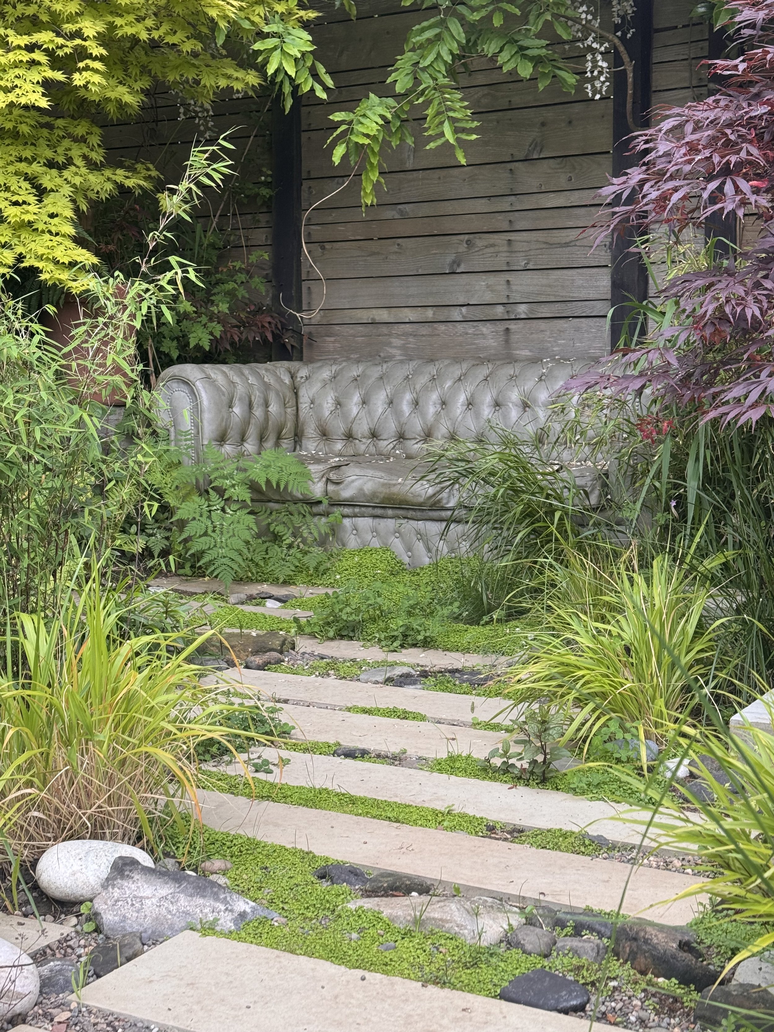 An outdoor scene featuring a concrete chesterfield sofa surrounded by green plants, bushes, and trees with a wooden fence in the background.