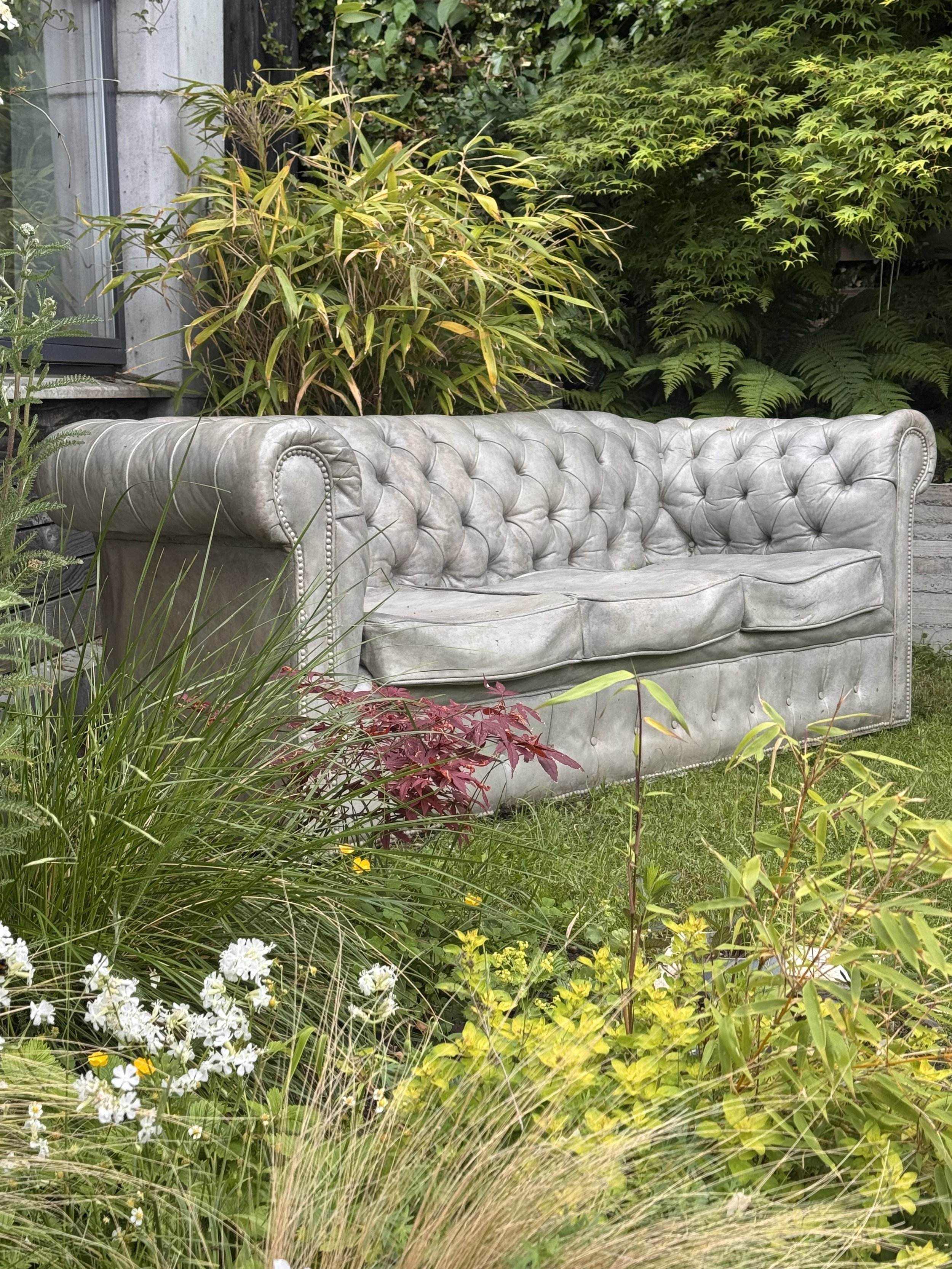 An outdoor scene with a concrete chesterfield sofa surrounded by green plants and flowers in a garden.