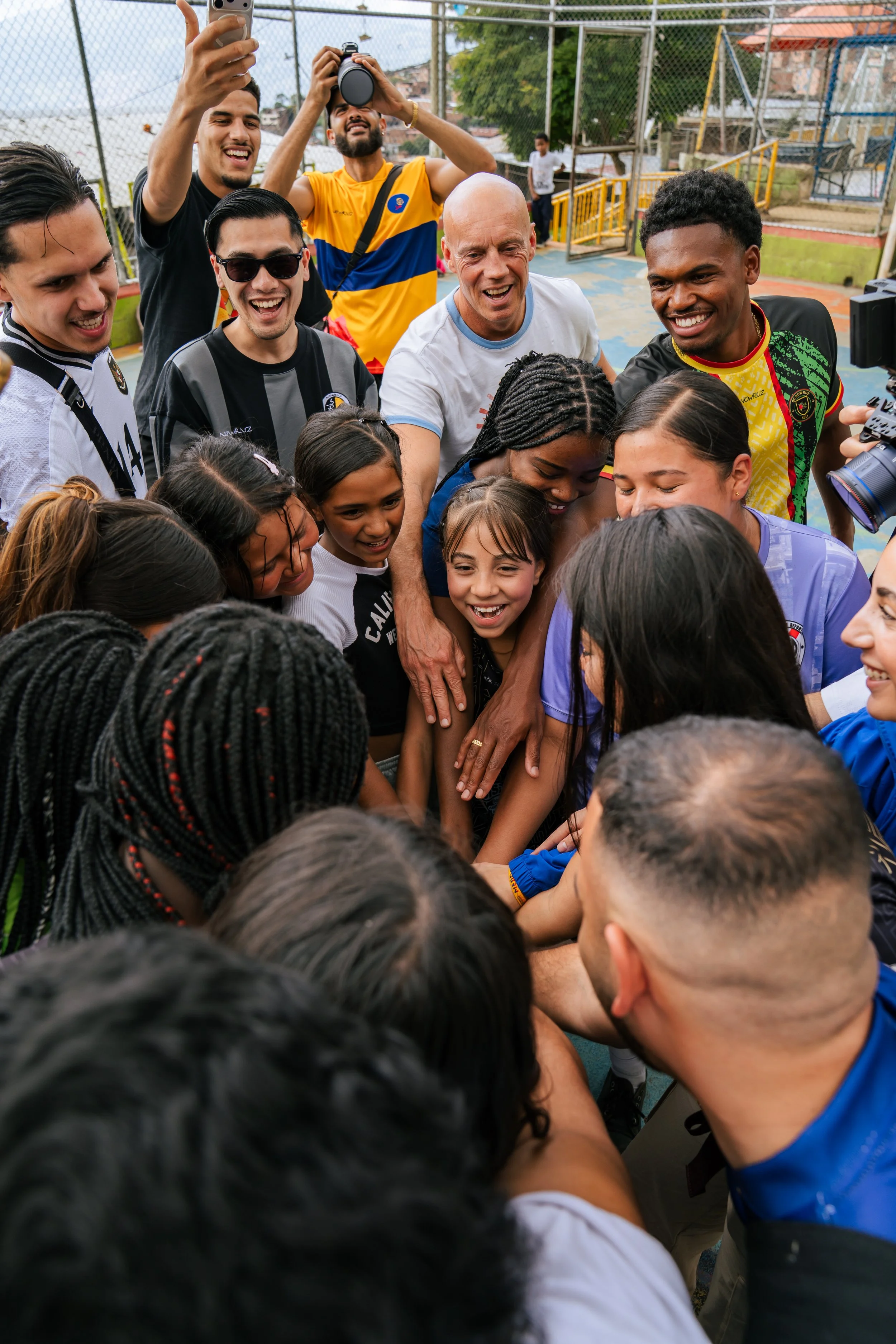 A diverse group of children and adults on a sports court, huddled together with their hands stacked in the center, smiling and celebrating.