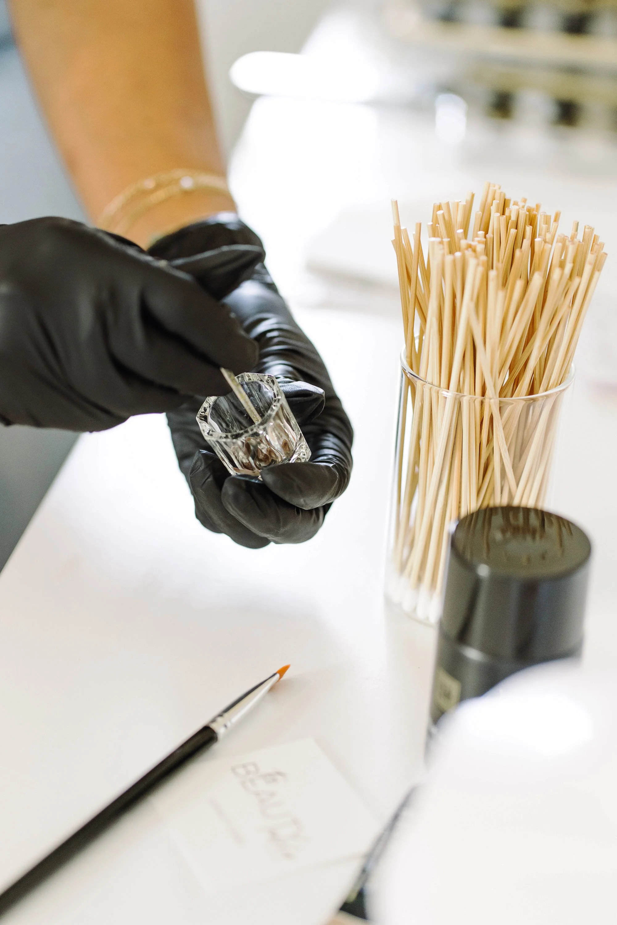 A person wearing black gloves holding a small glass container and adding liquid with a dropper. There is a container of uncooked spaghetti, a black container, a paintbrush, and a piece of paper on the white surface.