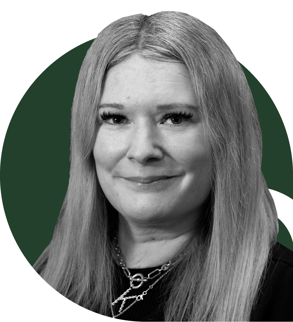 A woman with long blonde hair wearing a black top and a red blazer, standing indoors with a blurred background.