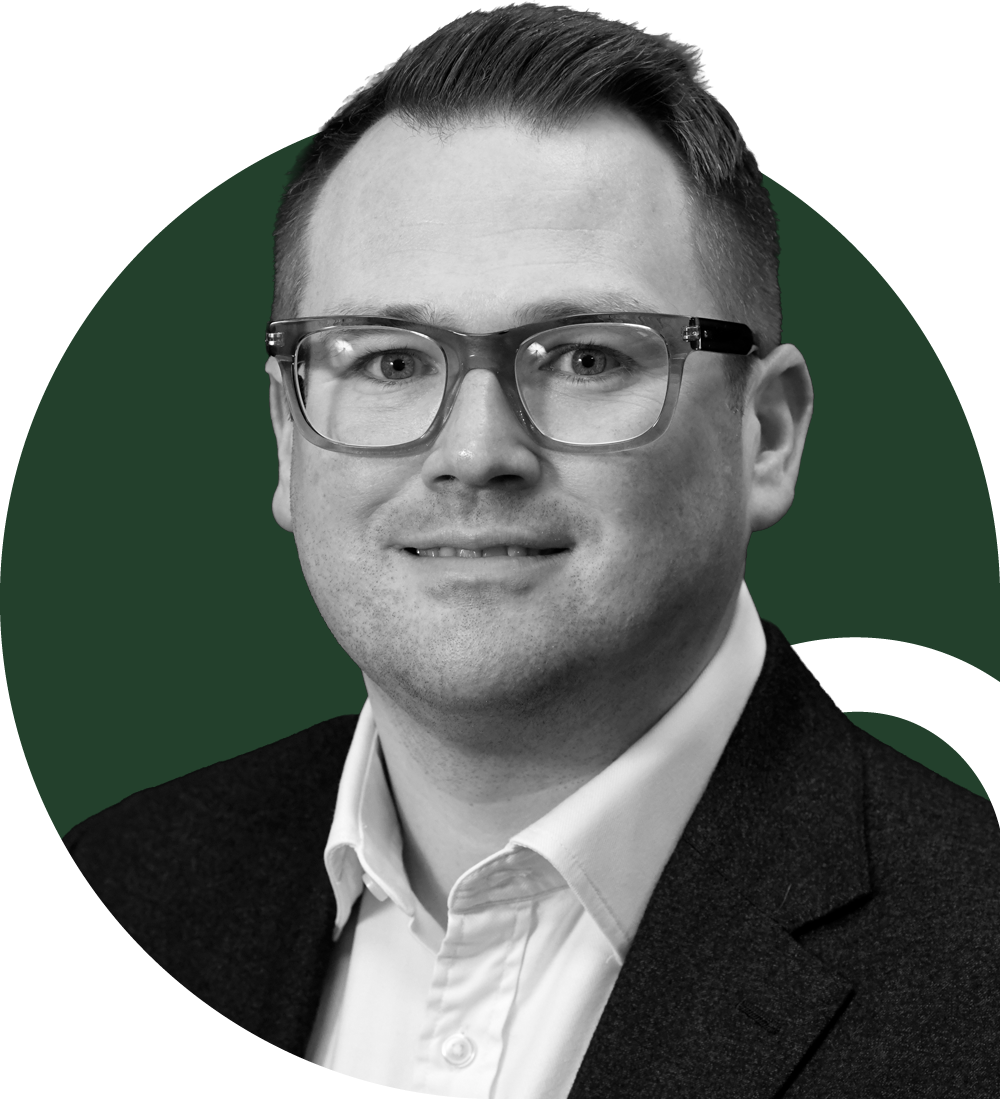 A man with glasses and short brown hair wearing a light blue dress shirt, looking at the camera in an indoor setting.