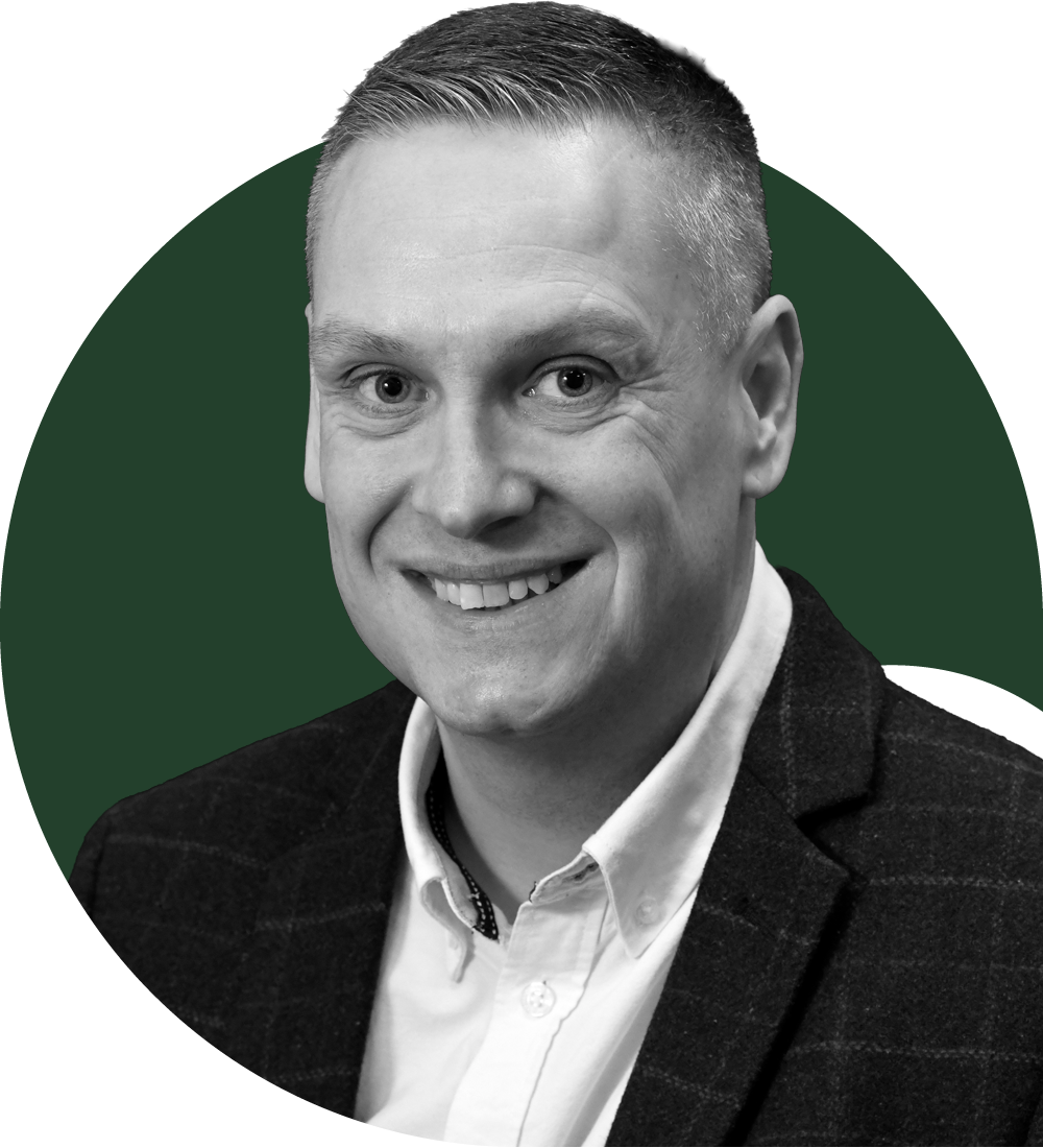 A man with short brown hair and blue eyes smiling, wearing a white shirt in an indoor setting with a blurred background.