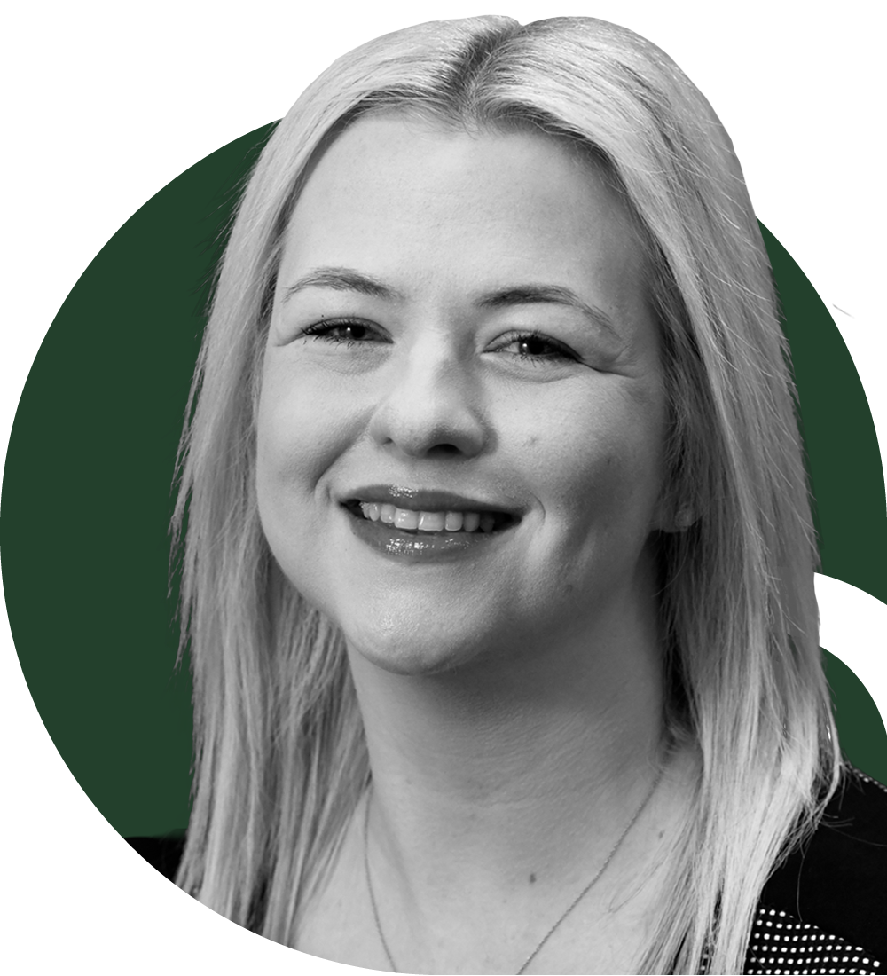 A blonde woman smiling in an indoor setting with large windows and a blurred background.