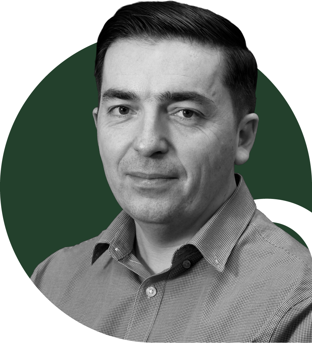 A man in a blue and white checkered shirt standing in a bright indoor space with large windows in the background.