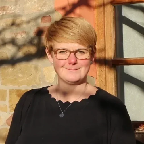 Eine Frau mit kurzen, blonden Haaren, Brille und schwarzem Oberteil steht vor einer Steinmauer und einem Fenster.