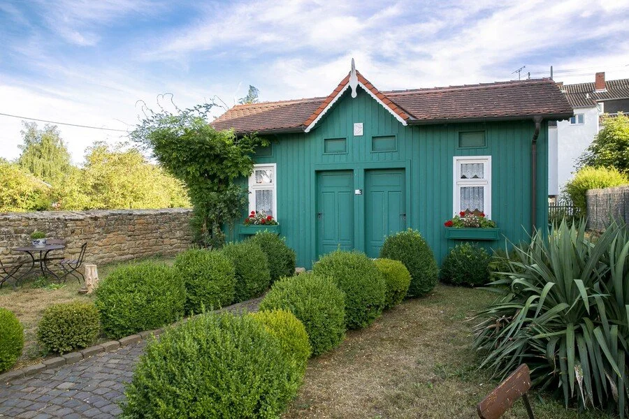 Ein grünes Haus mit rotem Dach, umgeben von einem Garten mit weißen Fensterrahmen, Balkonkästen mit roten Blumen, einem Kiesweg und verschiedenen grünen Pflanzen.
