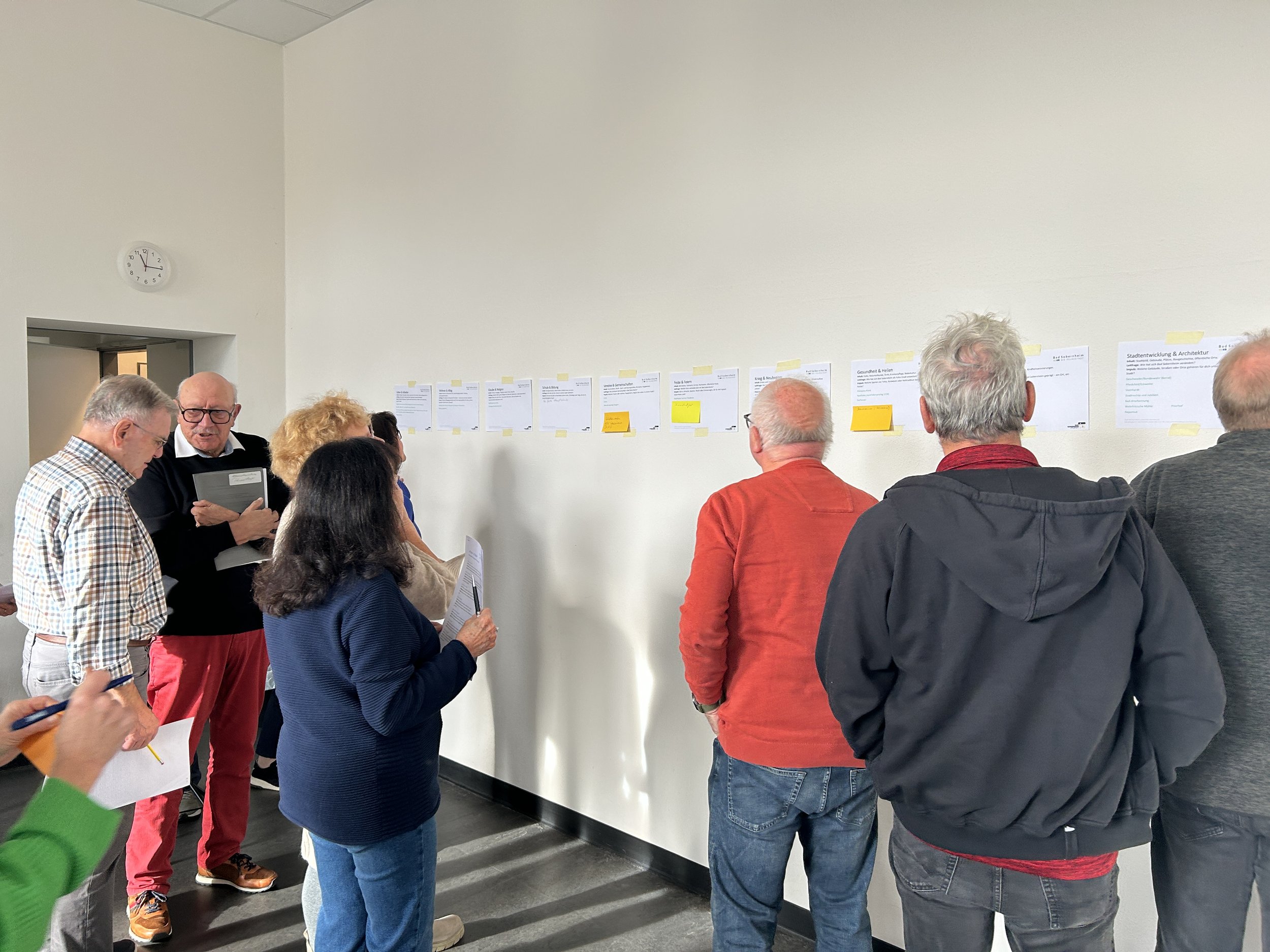 Menschen betrachten eine Wand mit Dokumenten, die mit Klebezetteln versehen sind, bei einer Besprechung in einem Raum.