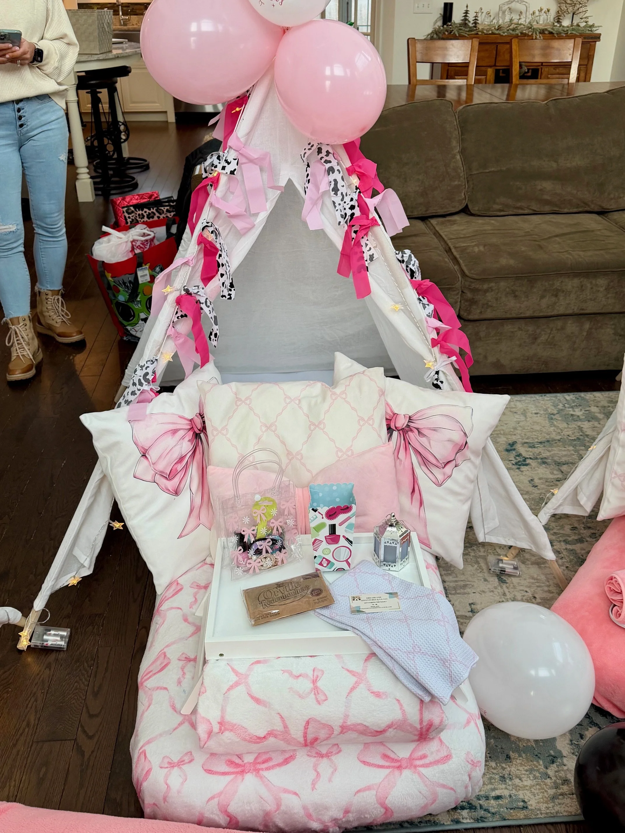 Pink bow-themed teepee sleepover setup for girls featuring a white indoor tent decorated with pink balloons, cow print and hot pink ribbon garlands, and soft twinkle lights. The bedding includes a pink bow blanket, plush pillows, and a tray with part