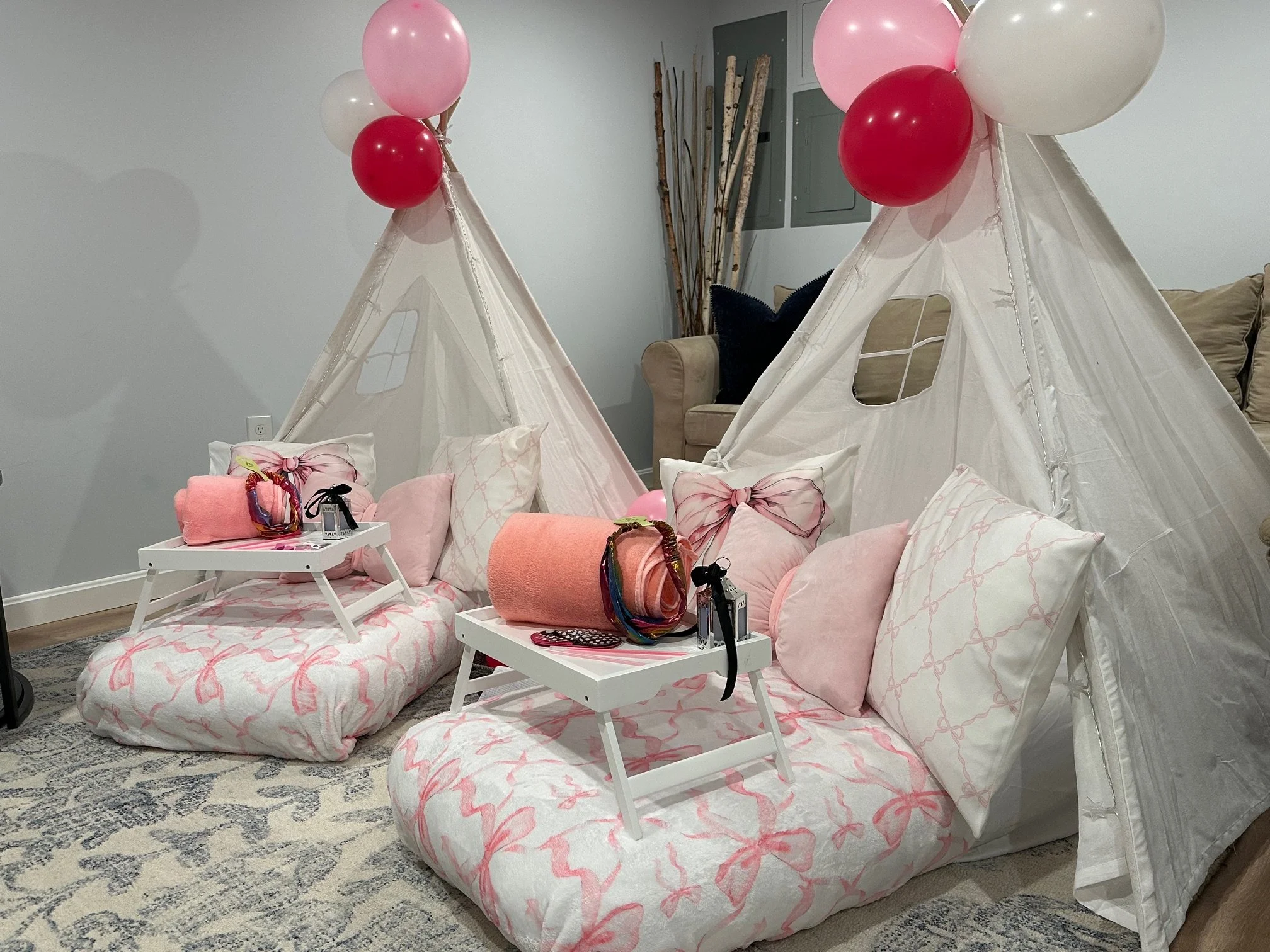 Pink bow-themed teepee sleepover setup for girls featuring a white indoor tent decorated with pink balloons. The beddings includes a pink bow blanket, plush pillows, and a tray with party favors, a lantern, and sleep masks.