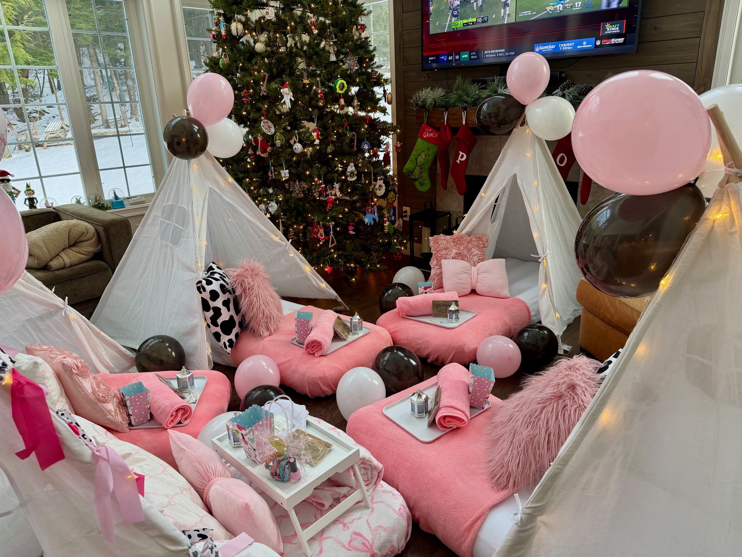 Stylish pink and black teepee sleepover setup for kids featuring white tents with fairy lights, blush pink bedding, fluffy pillows, and coordinated trays with spa-style details. Decor includes black, pink, and white balloons, fuzzy throw pillows, per