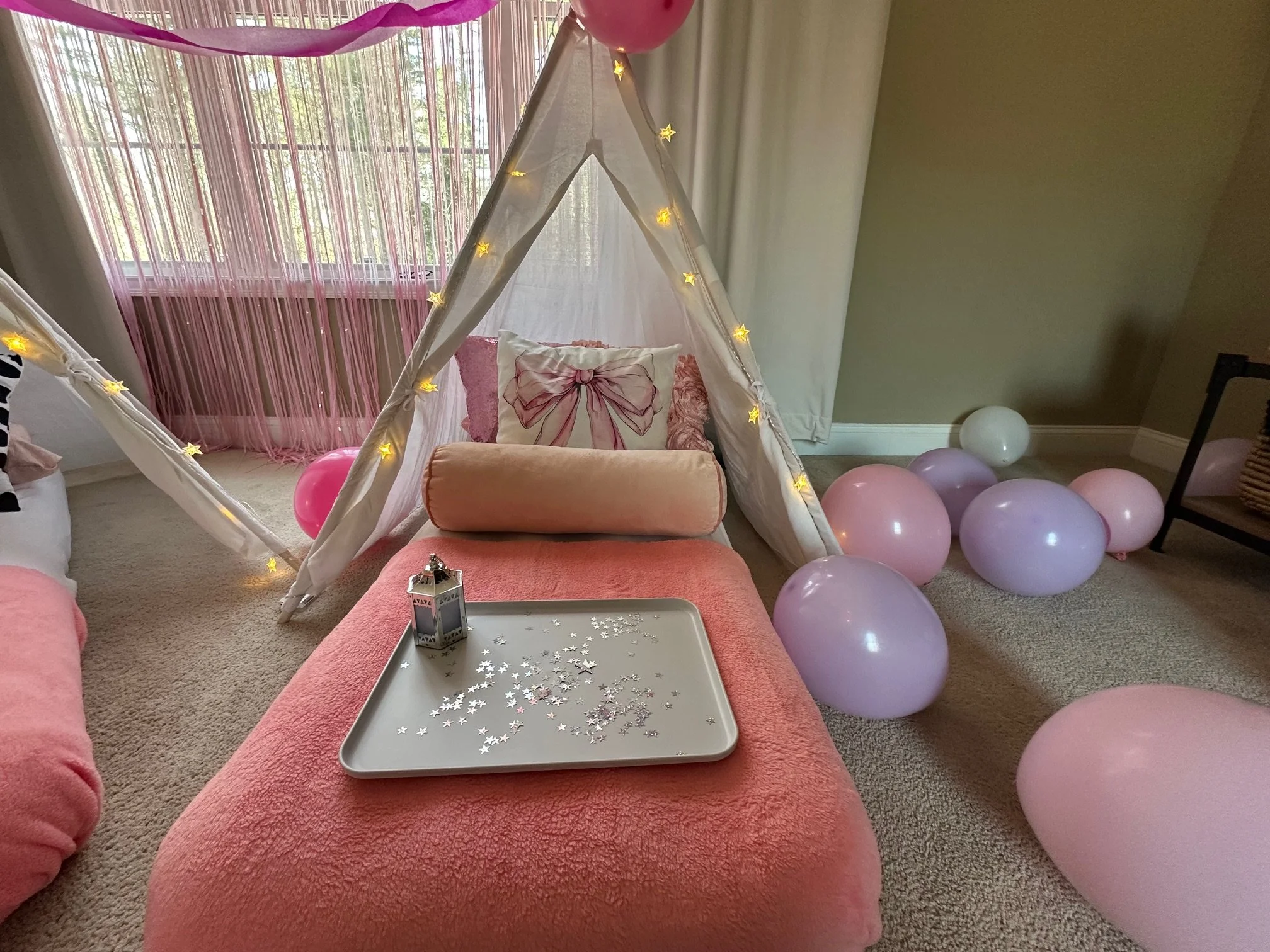 Pink and white teepee sleepover setup with twinkle lights, pink bedding, bow-themed pillows, and a tray with star confetti and a lantern, surrounded by pastel balloons.