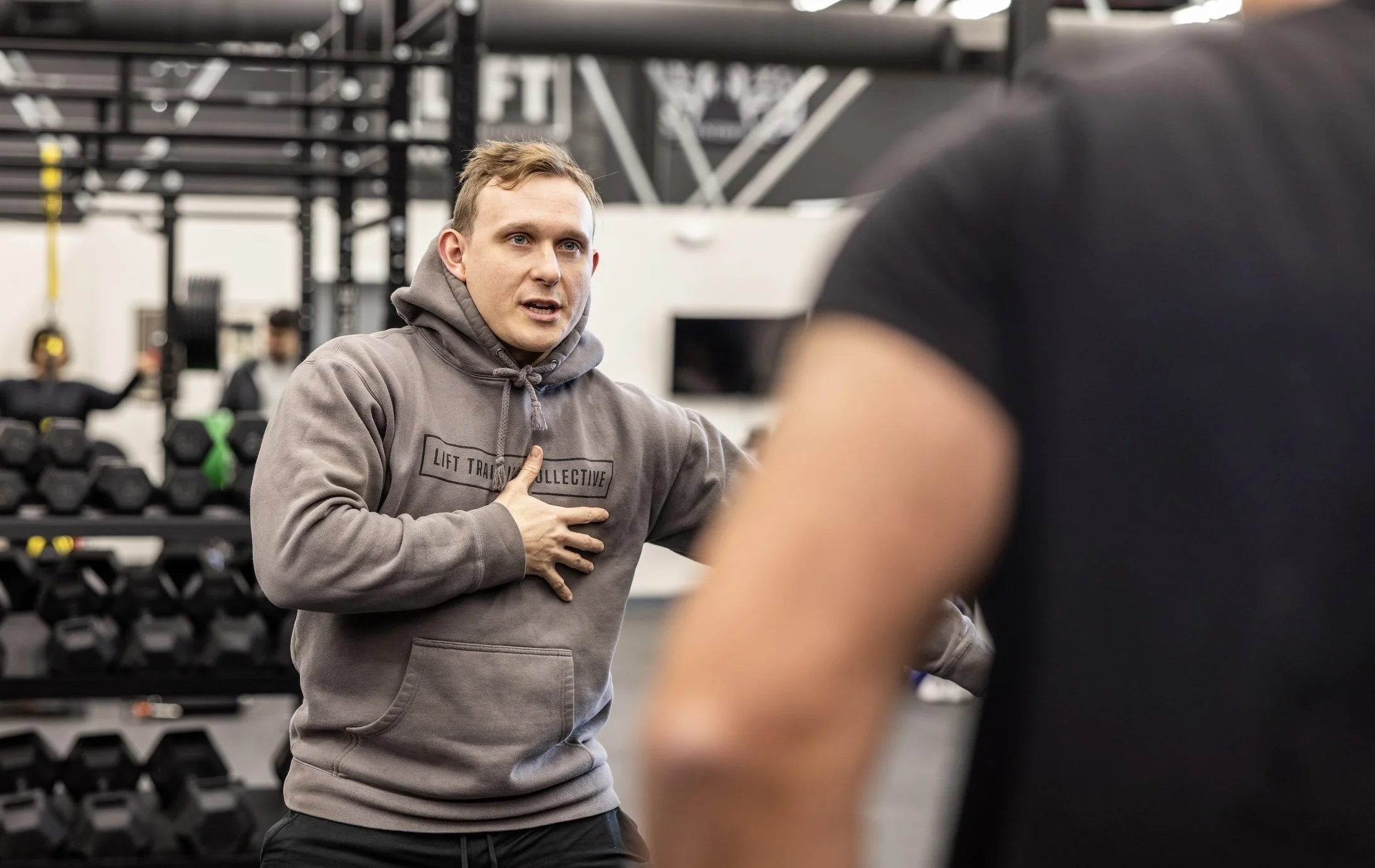 A trainer is speaking to a person in a gym, with weight racks and other gym equipment in the background.