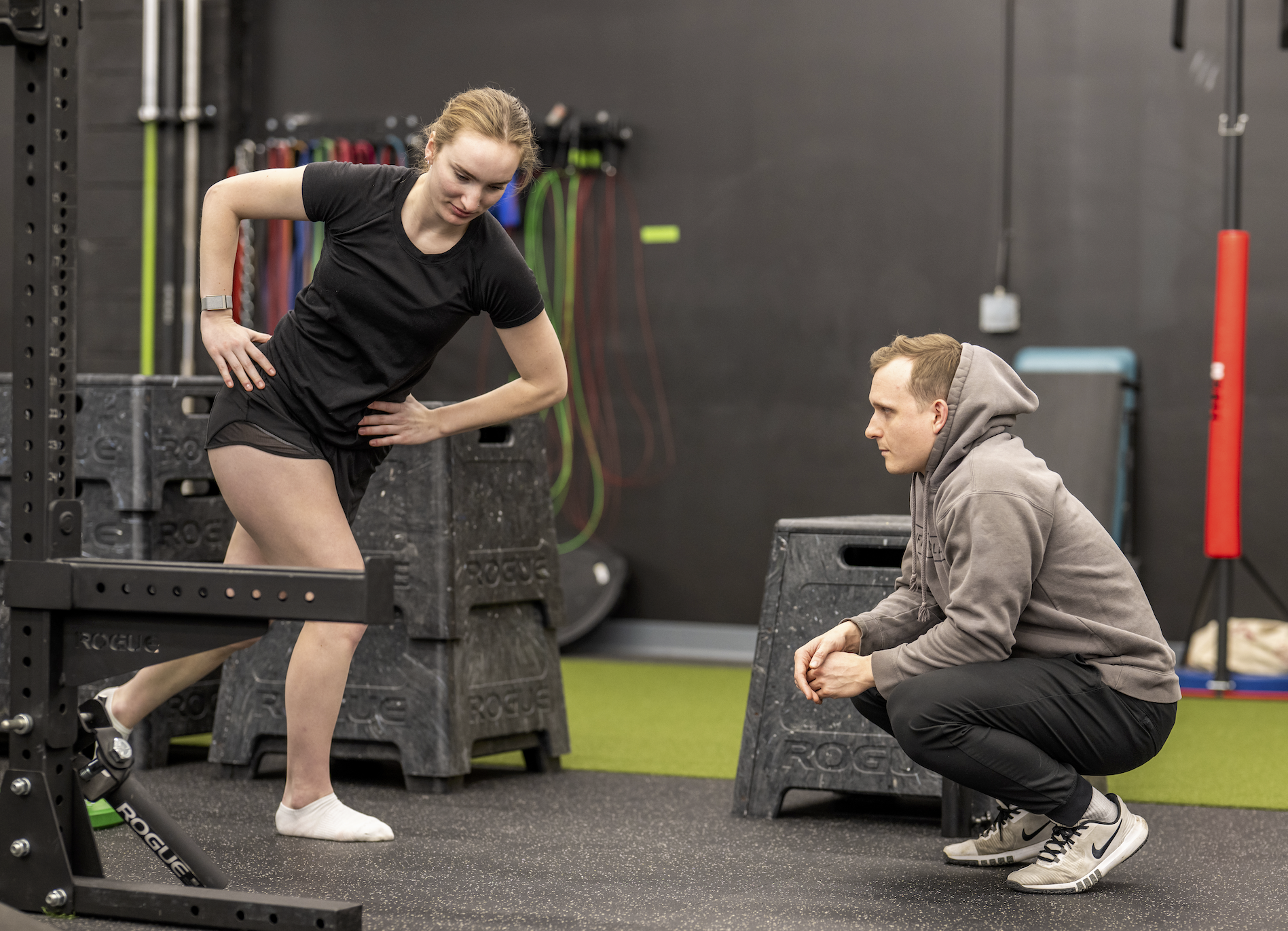 A woman in athletic clothing performing a workout with a weighted sled, while a man in a gray hoodie and black pants crouches nearby, observing or coaching in a gym with gym equipment and resistance bands in the background.