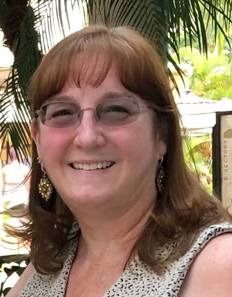 A smiling woman with brown hair and glasses, wearing large earrings and a sleeveless top, standing outdoors with lush tropical plants in the background.