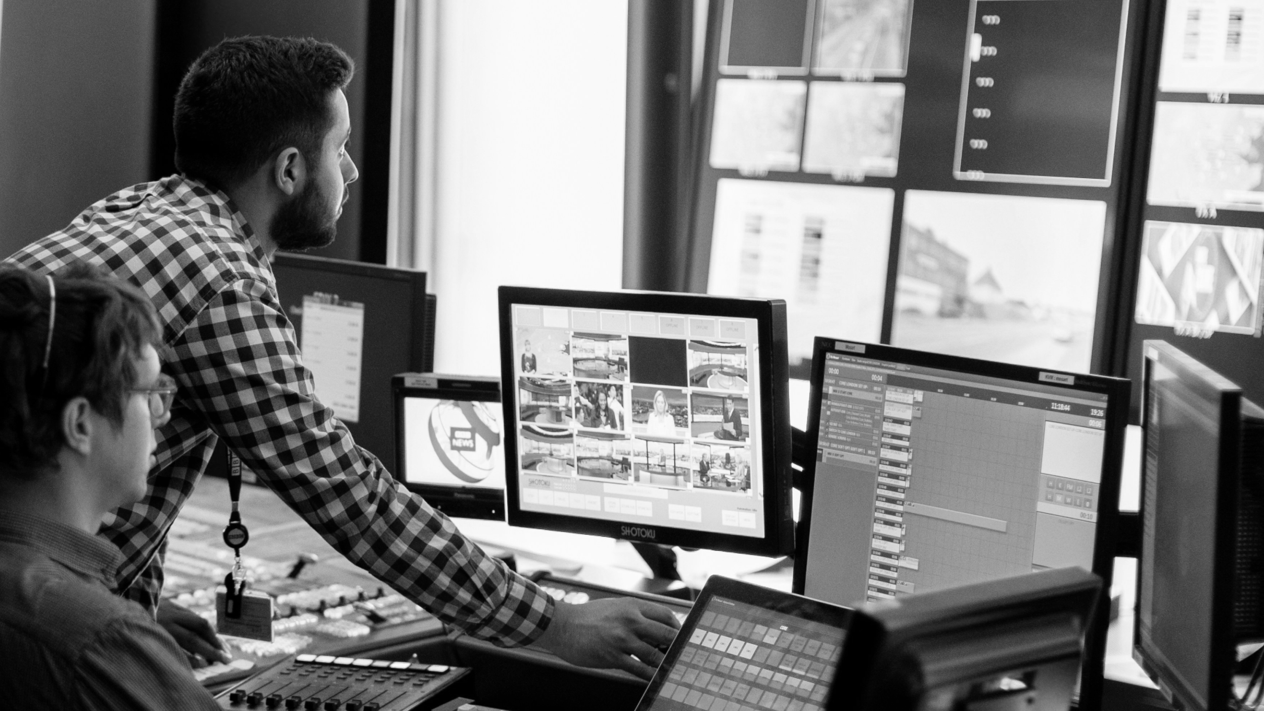 Two men working in a control room with multiple monitors displaying news and video production content.