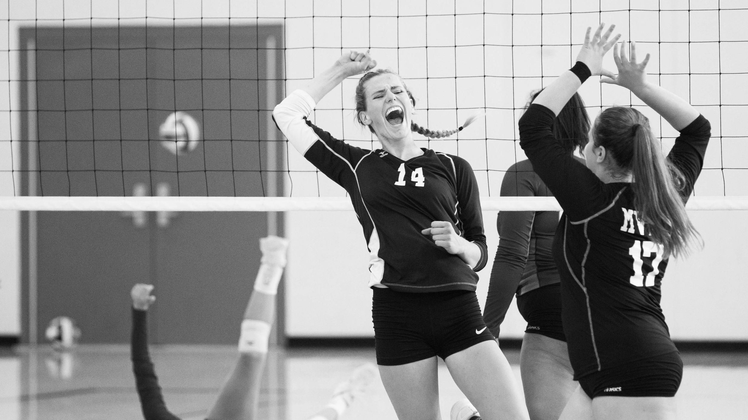 Black and white photo of volleyball players celebrating on the court. One player with number 14 is smiling and fist-pumping, while another with number 17 has her hands raised in the air. An opposing player is on the ground with her arm raised.