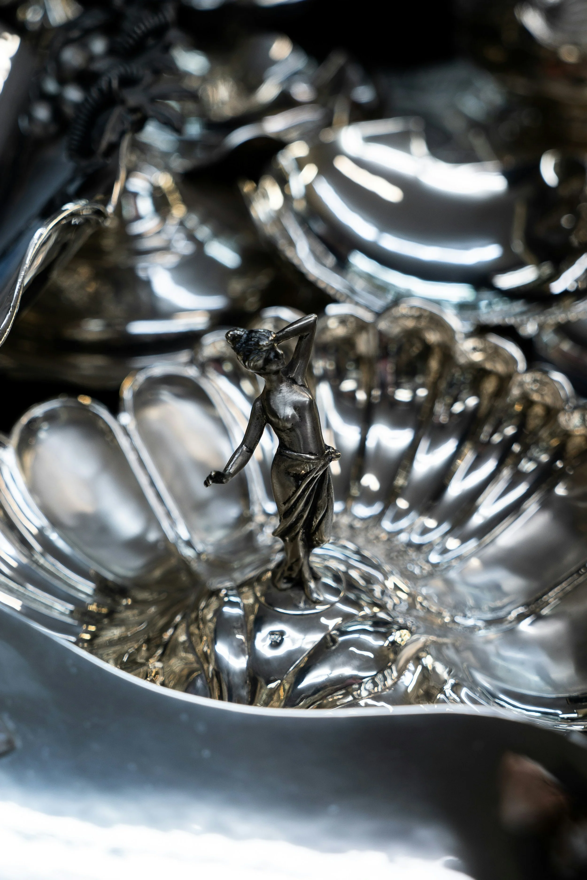 Close-up of a decorative silver sculpture with a small female figure standing on a shiny, reflective surface surrounded by various silver objects.