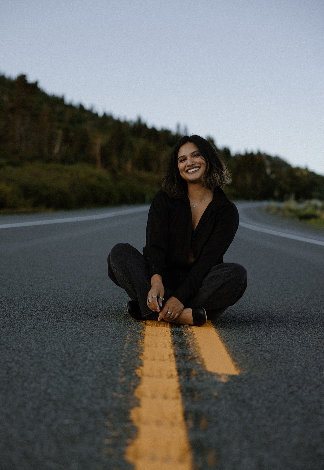 A woman sitting cross-legged on a deserted road with a yellow center line, smiling at the camera, with trees in the background under a clear sky.