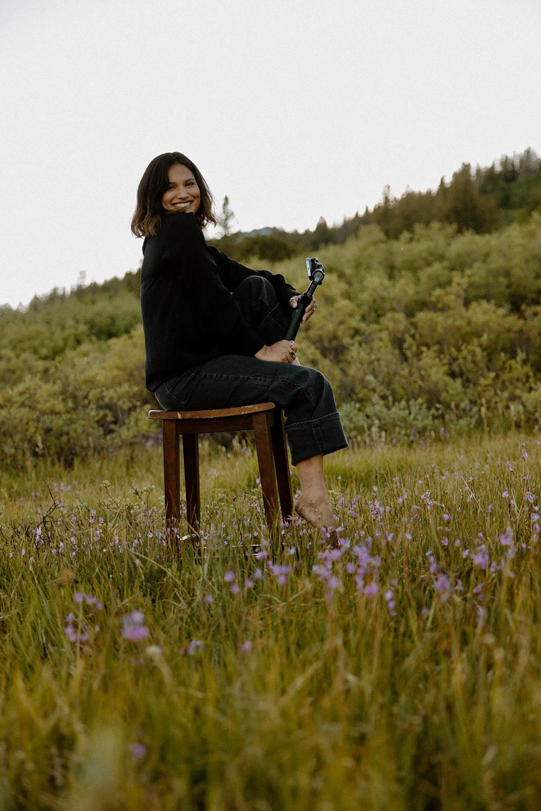A woman sitting on a wooden stool in a field of purple flowers, holding a selfie stick, smiling, with green hills in the background.
