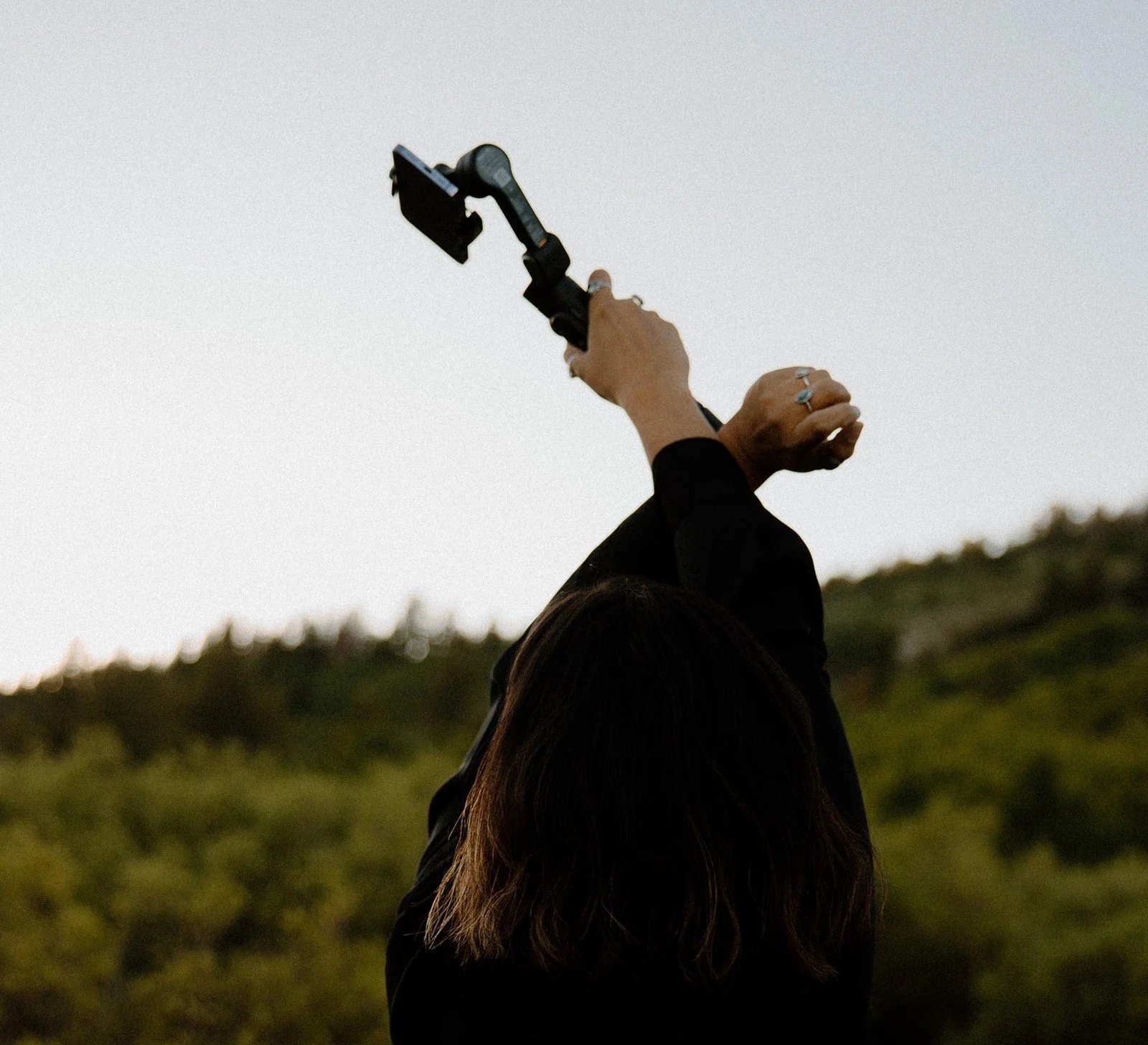 Person holding a black camera or phone with a stabilizer or tripod, raised towards the sky during sunset in a natural outdoor setting.