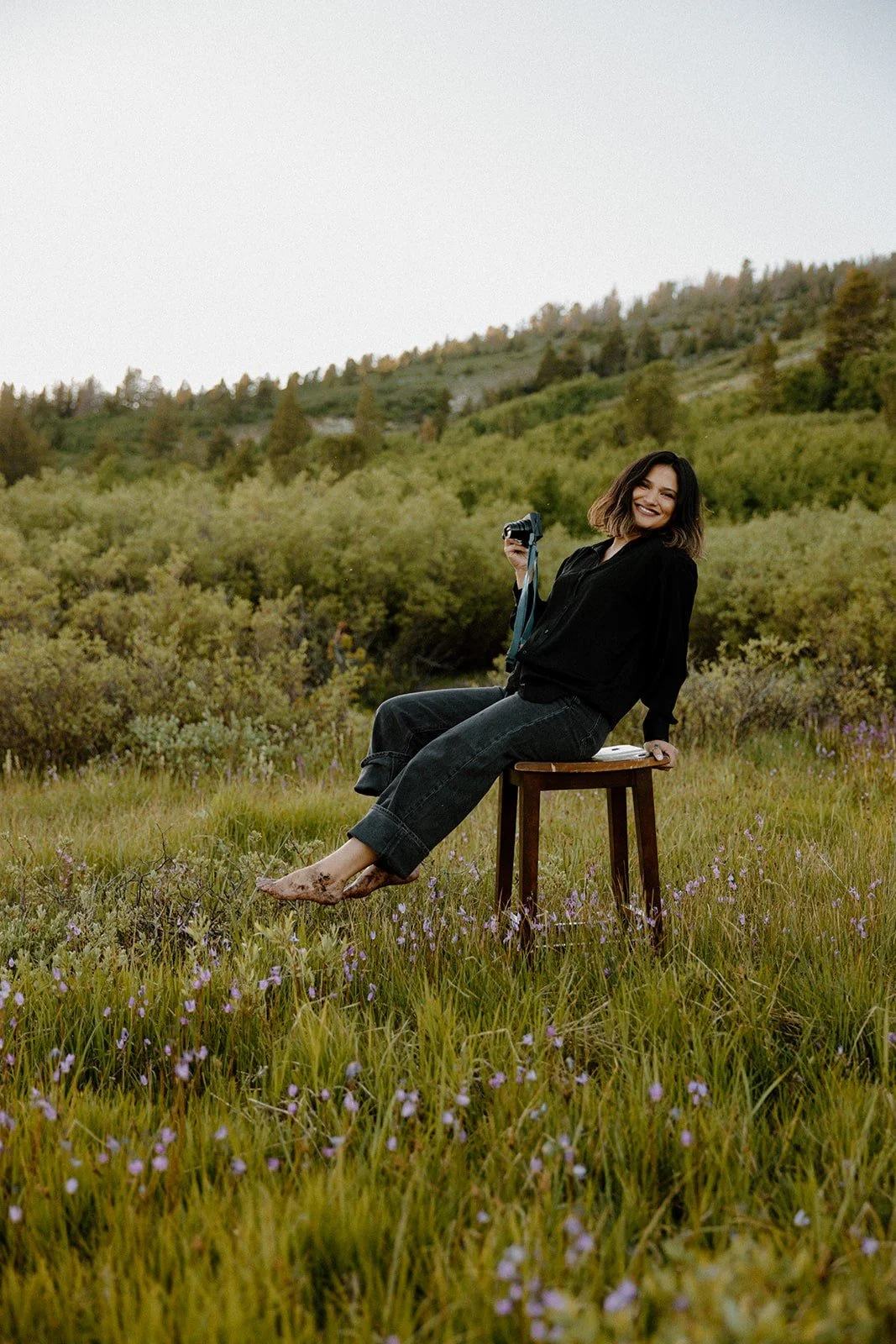 A woman sitting on a wooden stool in a grassy field with purple flowers, holding a camera, smiling, with a forested hill in the background.