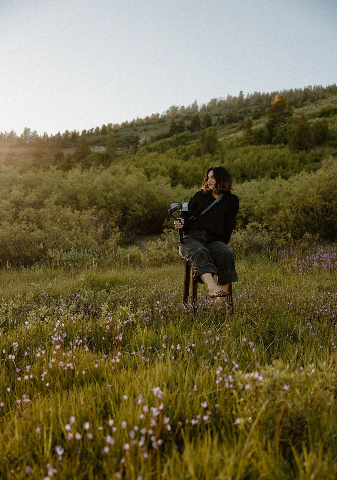 A woman sitting on a stool in a grassy field with purple flowers, holding a camera on a gimbal, during sunset with forested hills in the background.