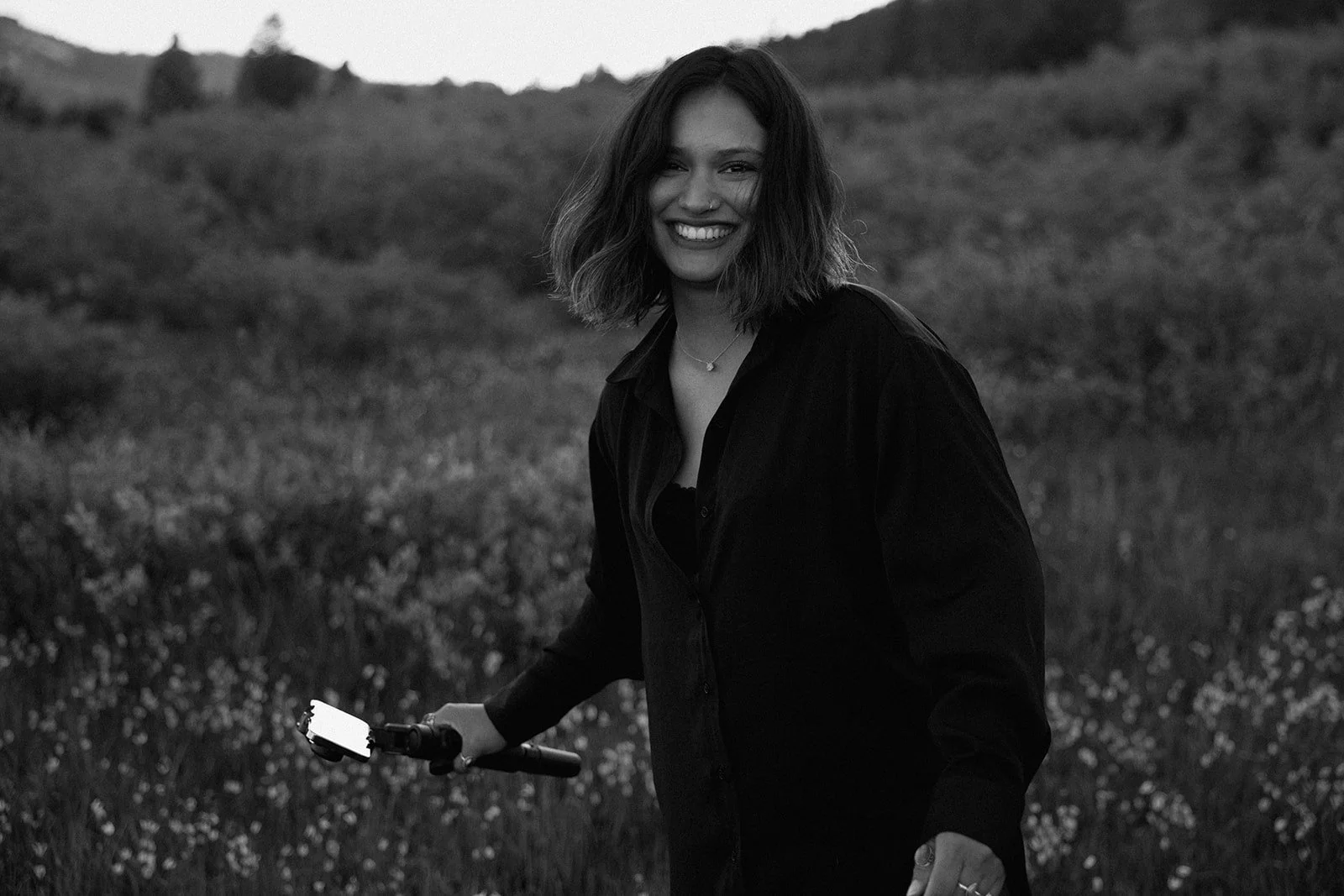 Black and white photo of a smiling woman with shoulder-length hair outdoors, holding a scooter in a grassy field with hills in the background.