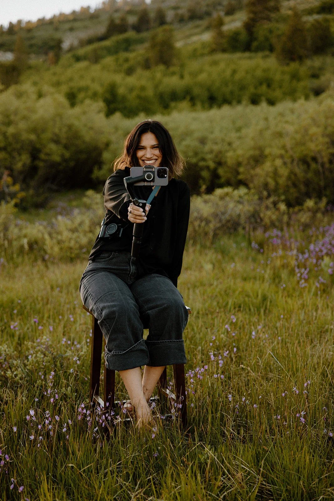 A woman taking a selfie outdoors in a grassy field with purple flowers, wearing black clothing, sitting on a stool, with a scenic, green hillside in the background.