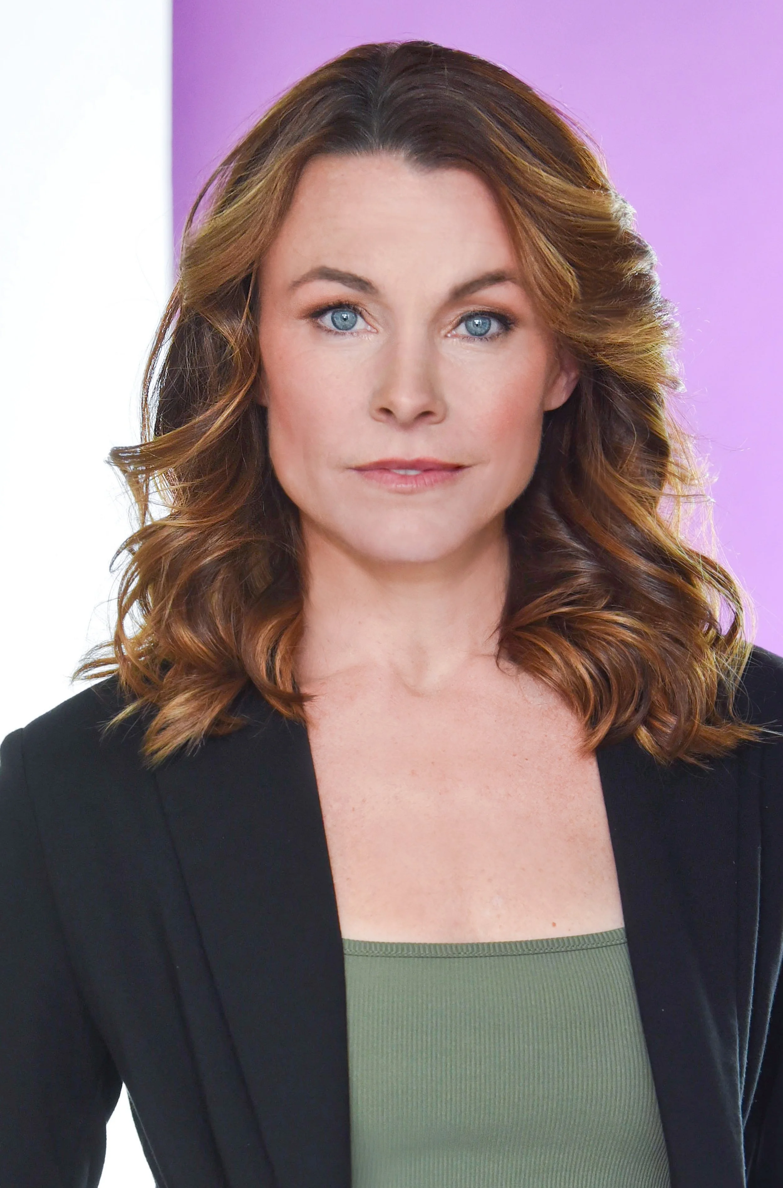 A woman with wavy brown hair and blue eyes wearing a black blazer and green top, standing in front of a purple and white background.