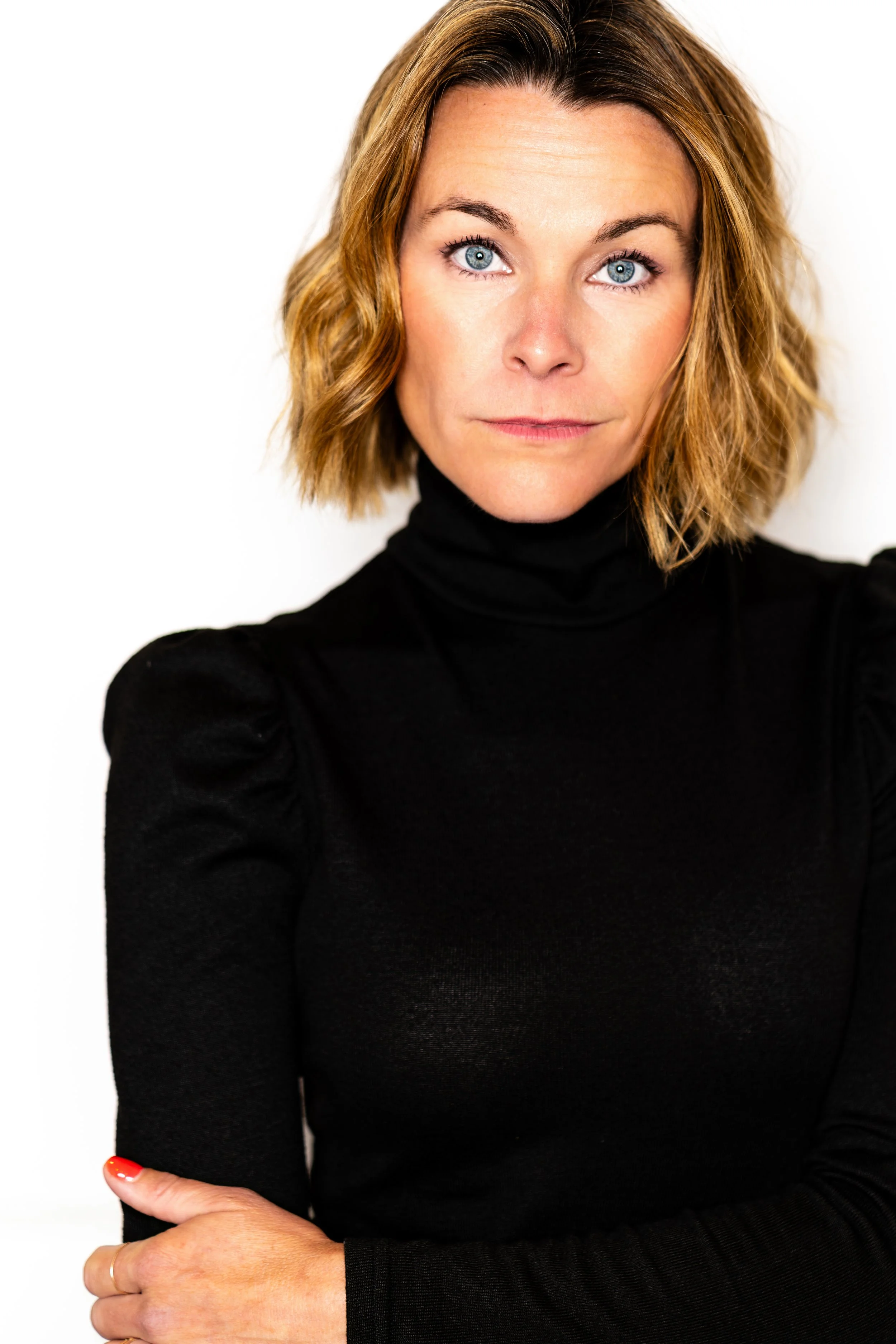 A woman with short wavy blonde hair, blue eyes, wearing a black turtleneck, standing against a white background.