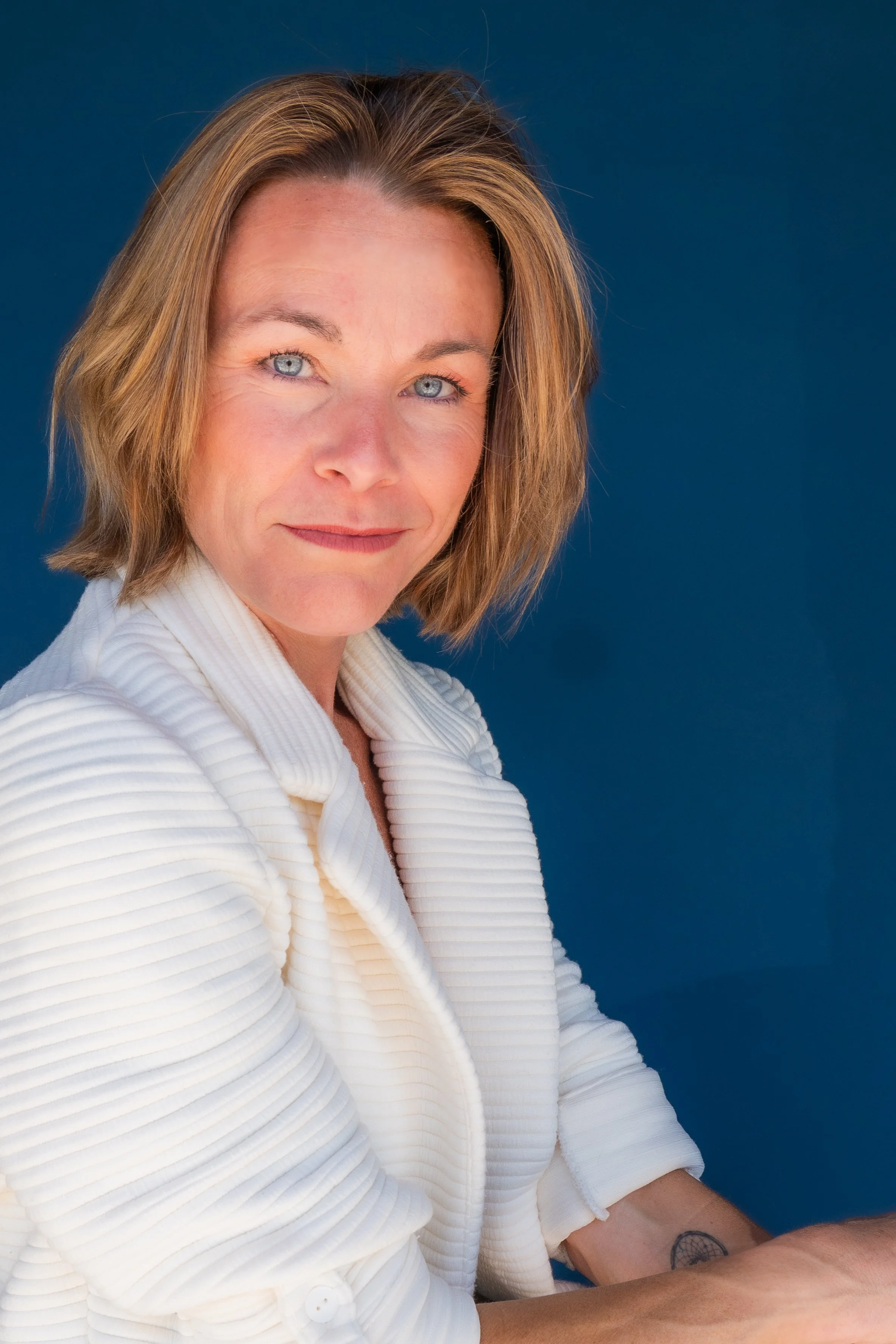 A woman with shoulder-length brown hair, wearing a white textured blazer, looking at the camera with a slight smile against a blue background.