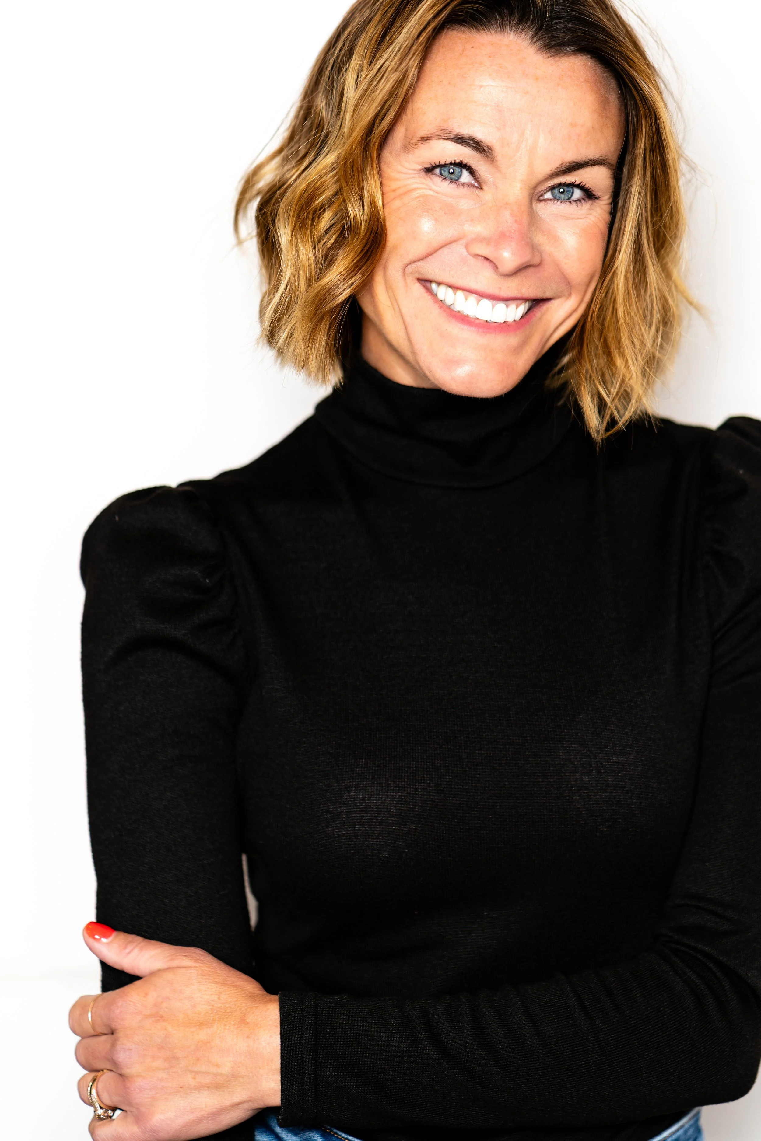 A smiling woman with blonde hair and blue eyes wearing a black turtleneck and posing with crossed arms against a white background.