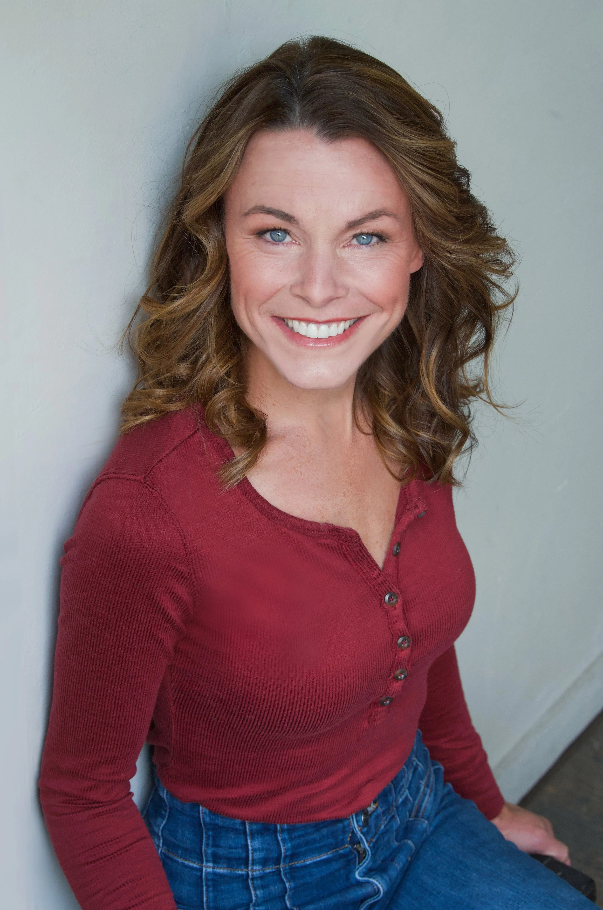 A woman with wavy brown hair smiling, wearing a red long-sleeve shirt and blue jeans, sitting against a light-colored wall.