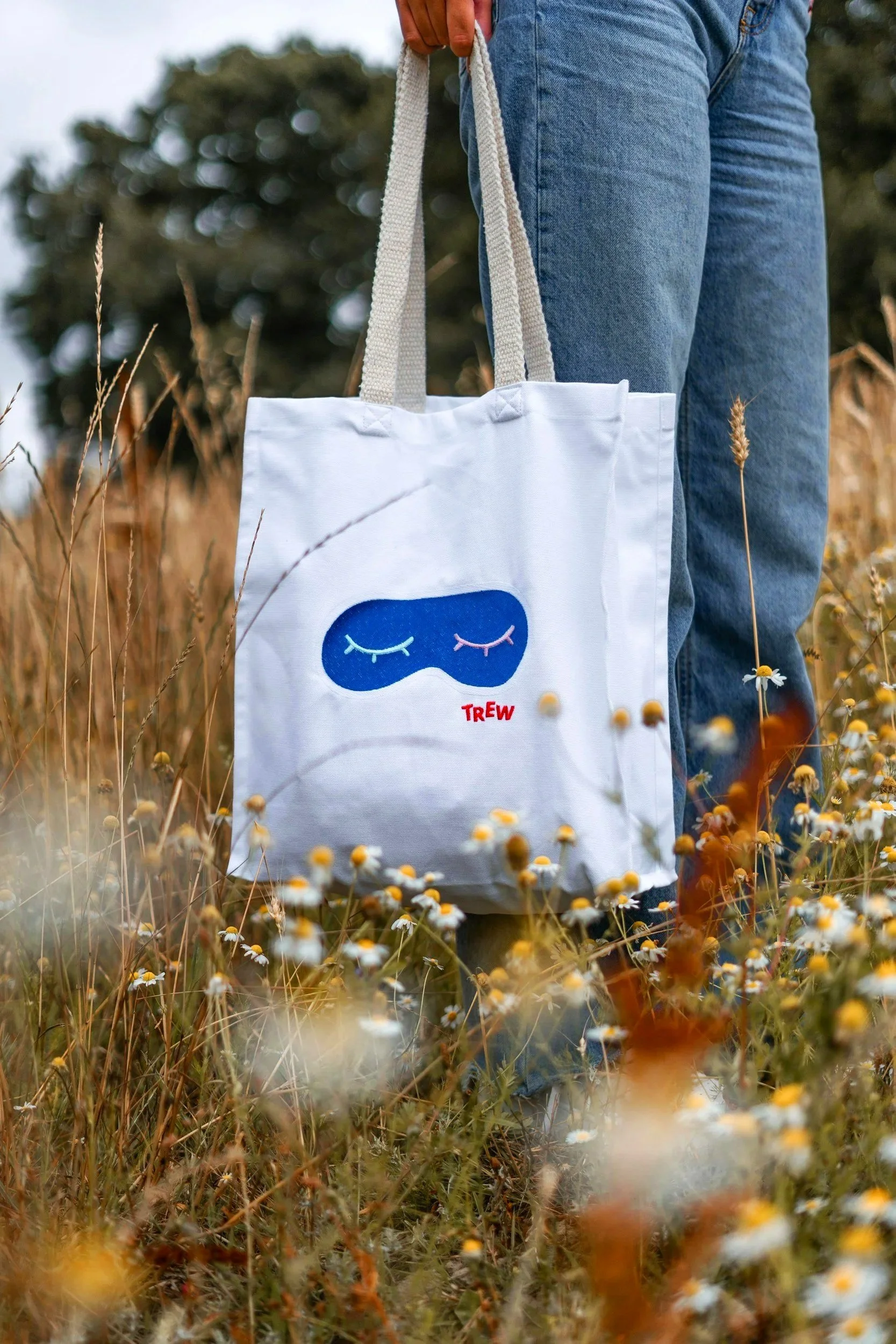 Person standing in a field of wildflowers holding a reusable tote bag with a sleeping mask face design and the word 'TREW' on it, wearing blue jeans.